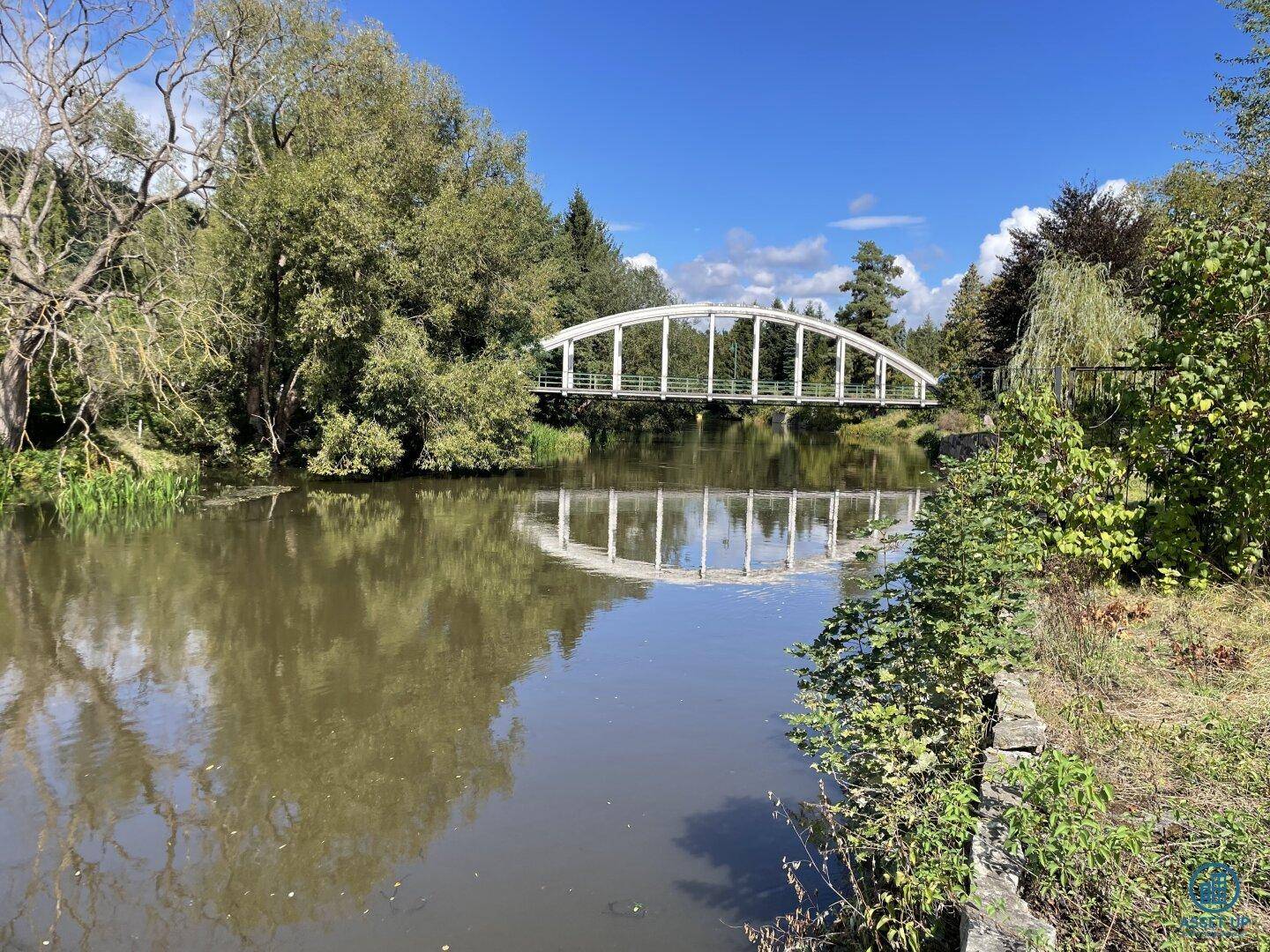 Idyllischer Fluss mit einer weißen Bogenbrücke, die sich im Wasser spiegelt, umgeben von Bäumen.