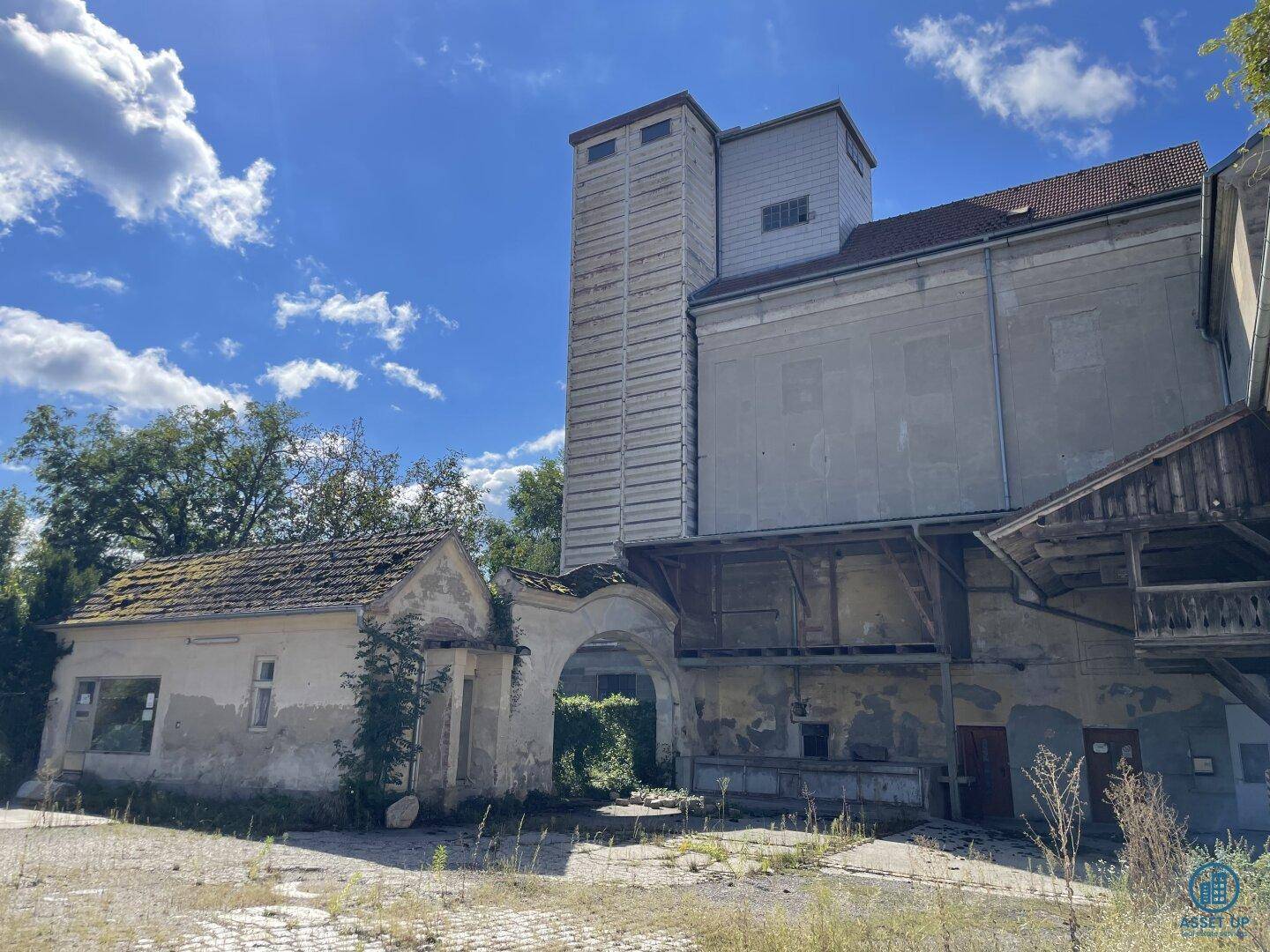 Historischer Gebäudekomplex mit einem kleinen Haus, Torbogen und einem hohen Turm unter bewölktem Himmel.