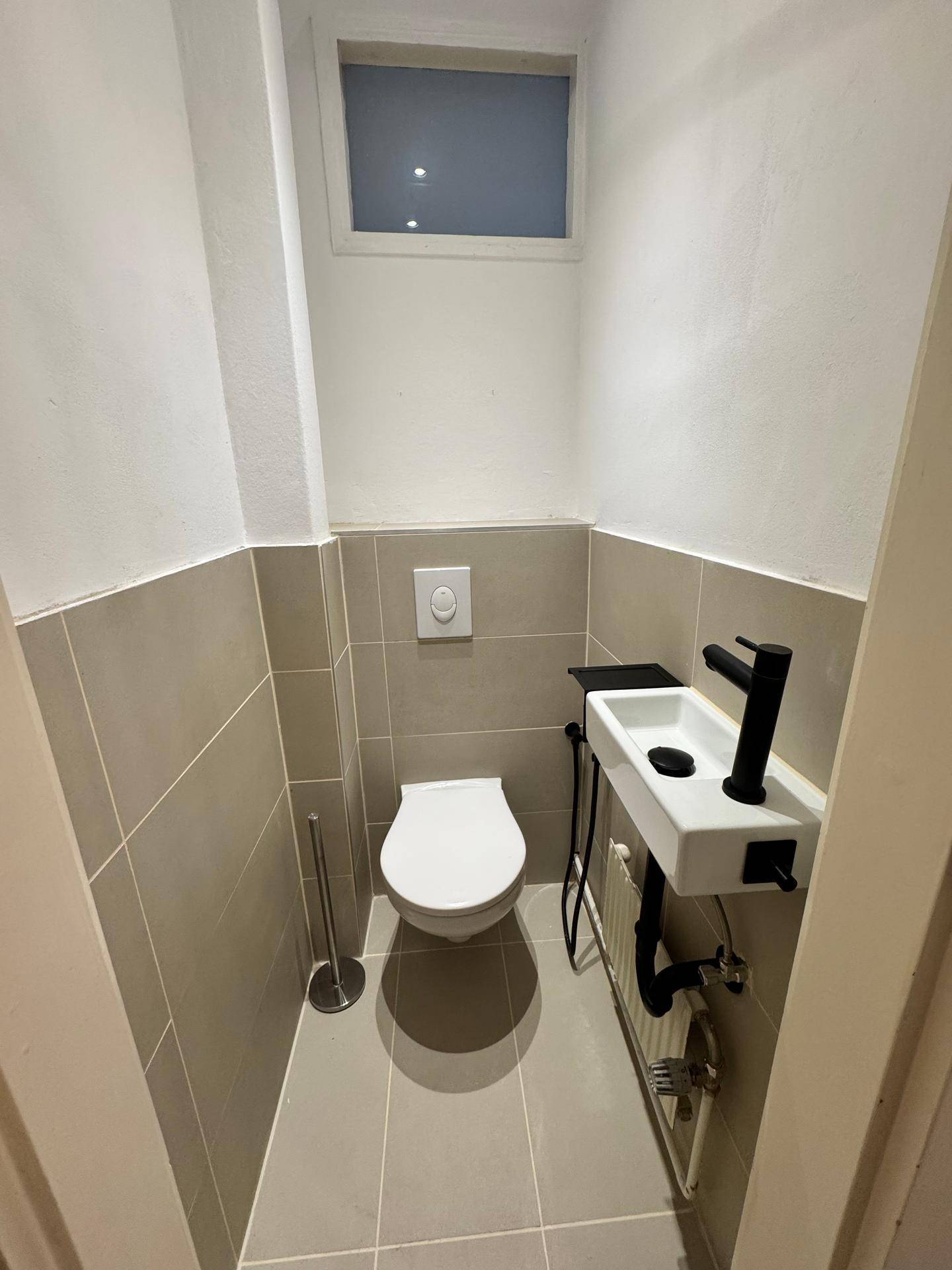 Gäste-WC mit wandhängender Toilette, kleinem Waschbecken und einem Fenster für natürliche Belüftung.