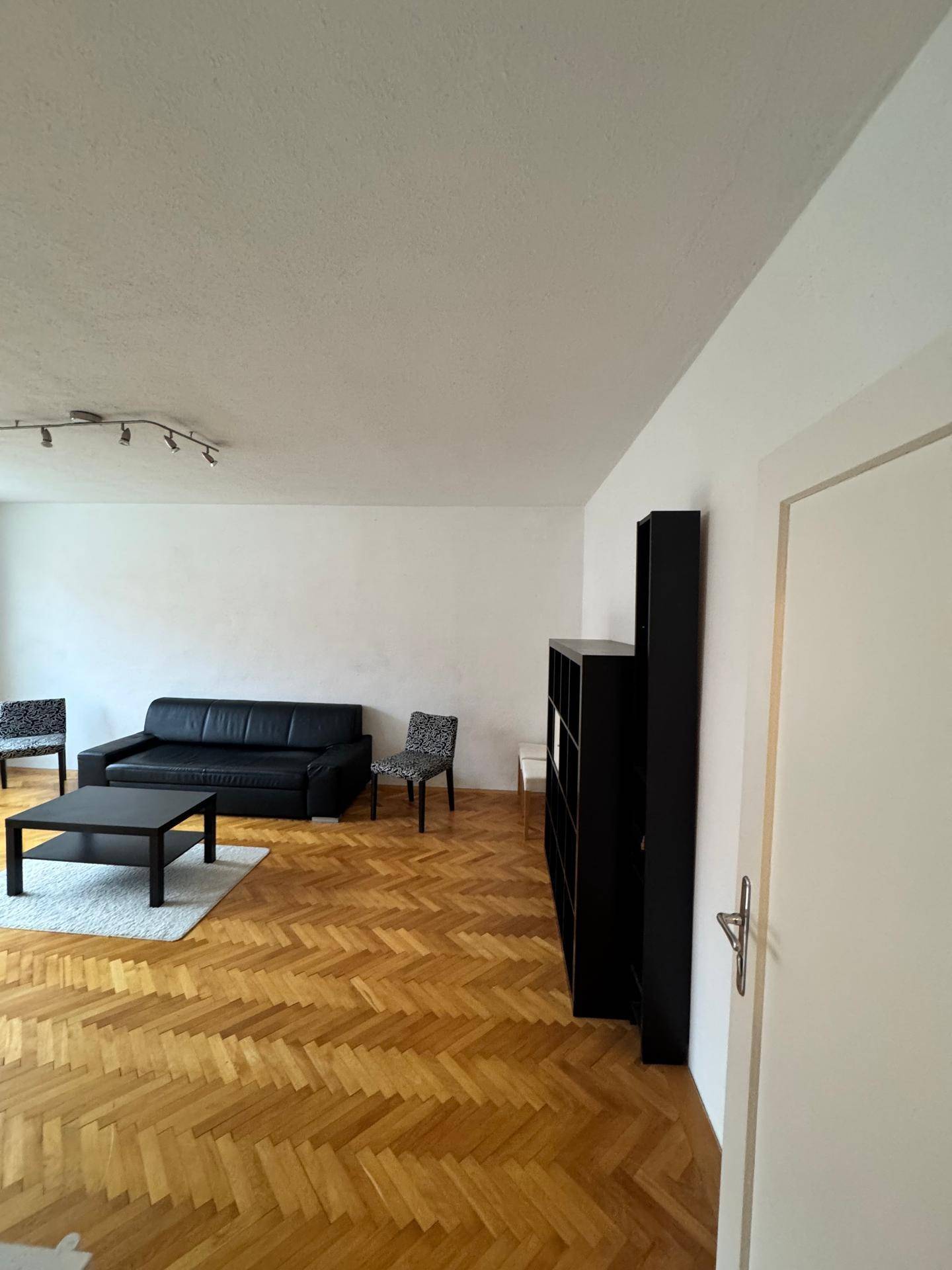 Wohnzimmer mit Fischgrätparkett, schwarzem Sofa und Bücherregal, das eine gemütliche Atmosphäre schafft.