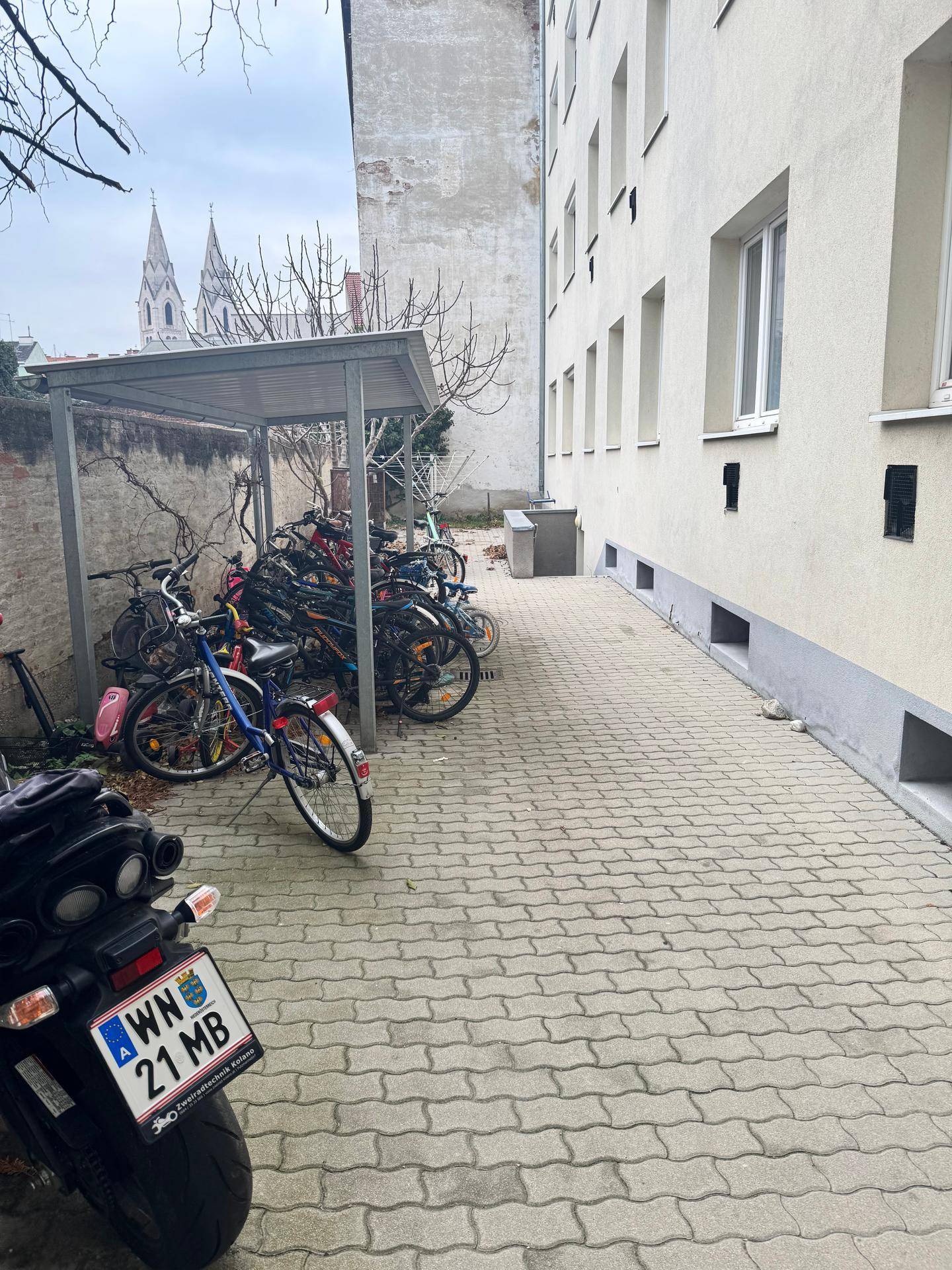Fahrradabstellplatz mit Überdachung neben einem Gebäude, im Hintergrund ist ein Kirchturm zu sehen.