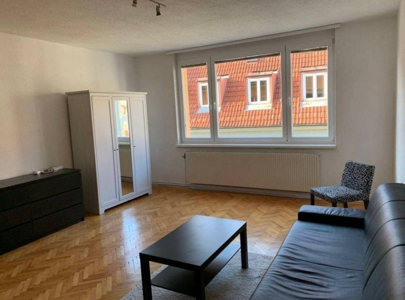 Geräumiges Wohnzimmer mit Holzparkett, weißem Kleiderschrank und Blick auf die umliegenden Dächer.