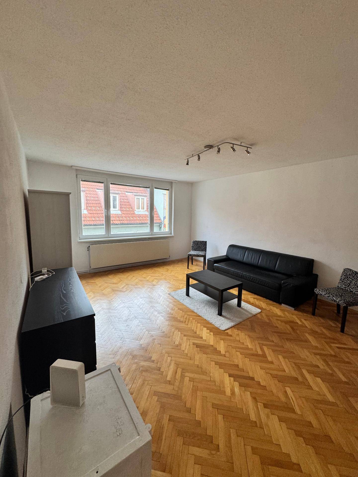 Helles Wohnzimmer mit Fischgrätparkett, schwarzem Sofa und Blick auf die Dächer durch ein großes Fenster.