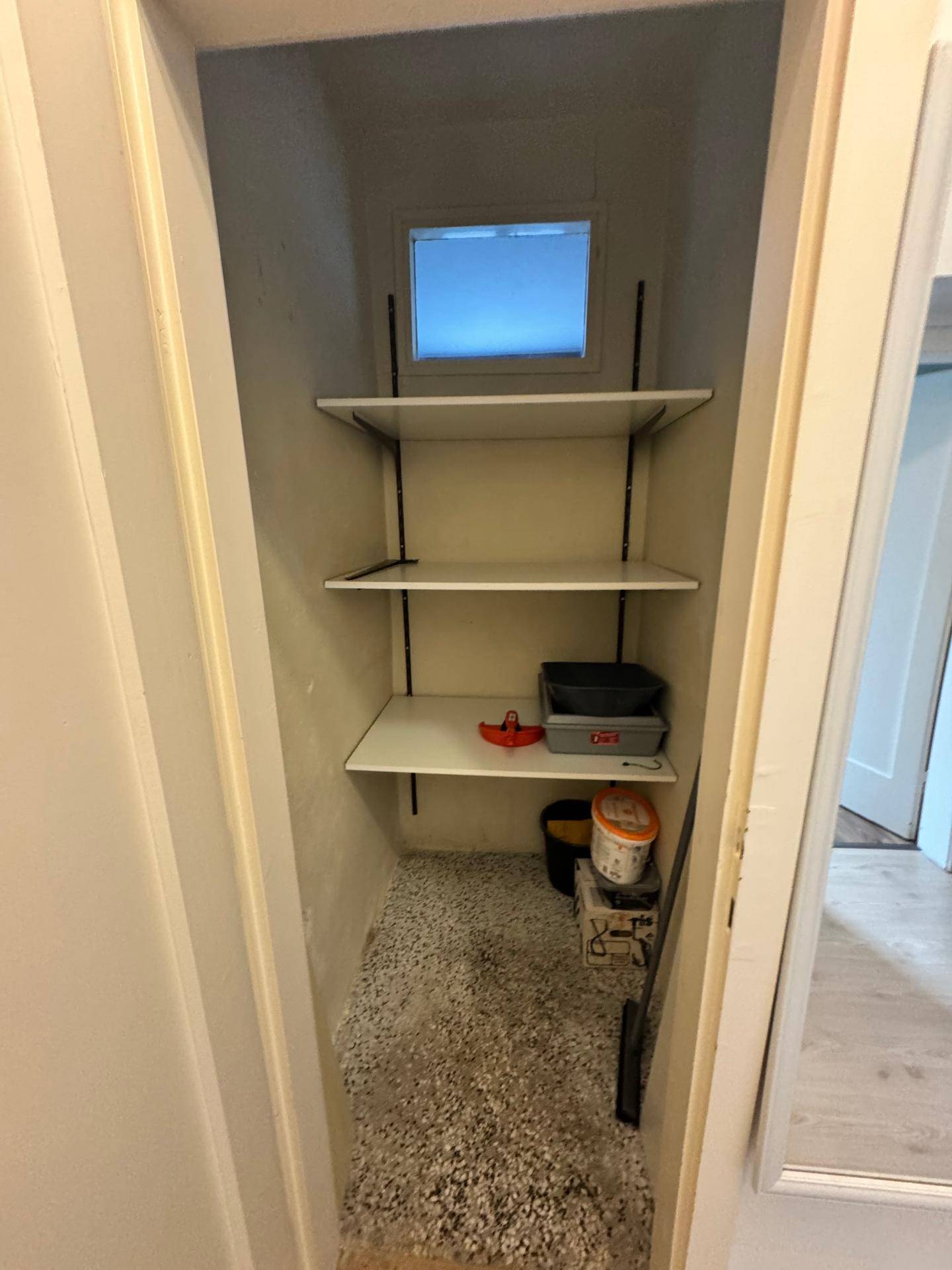 Kleiner Abstellraum mit Regalen, einem kleinen Fenster und einem Bodenbelag aus Kies.