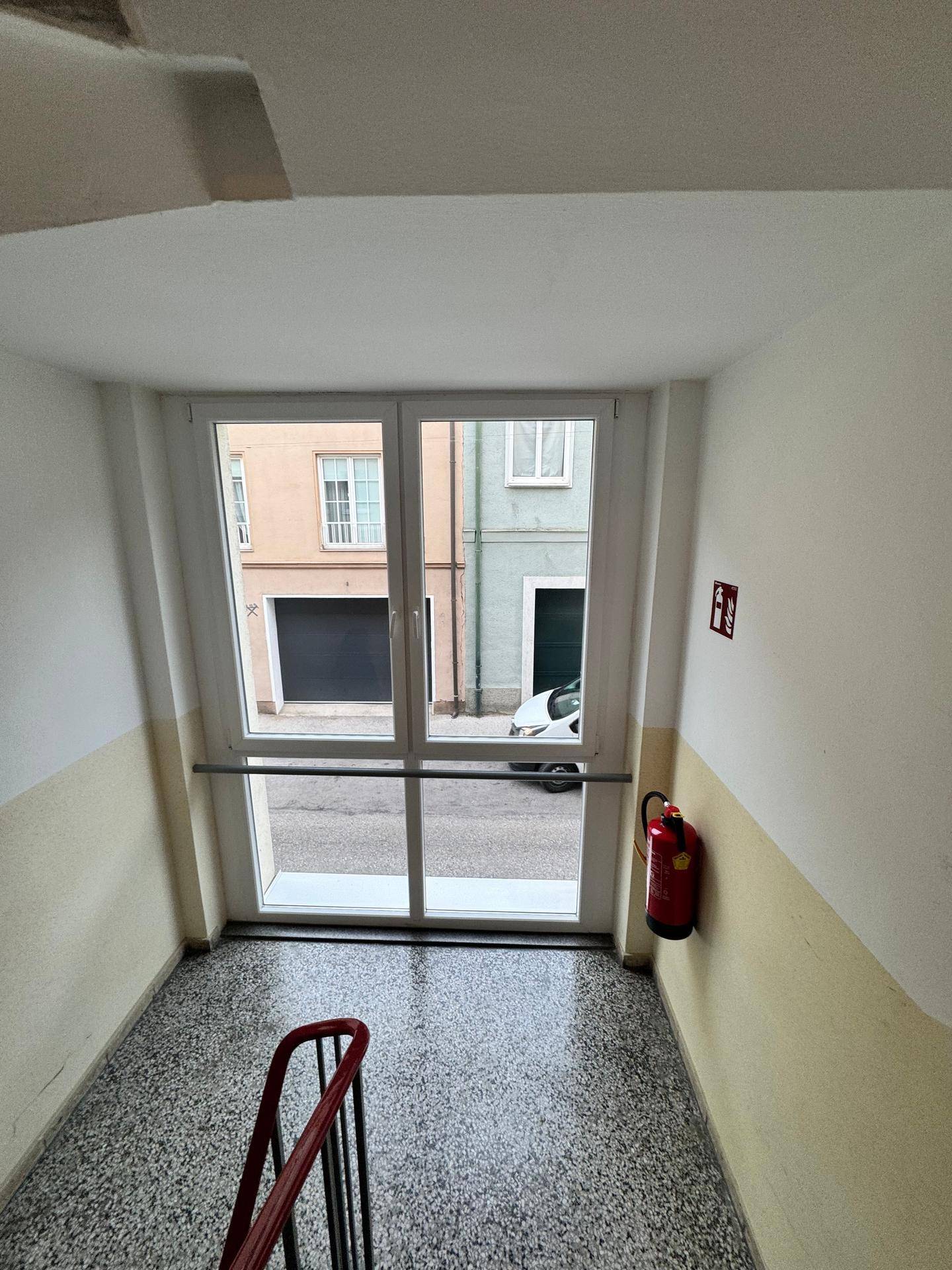 Treppenhaus mit Terrazzoboden, rotem Geländer und einem großen Fenster mit Blick auf die Straße.