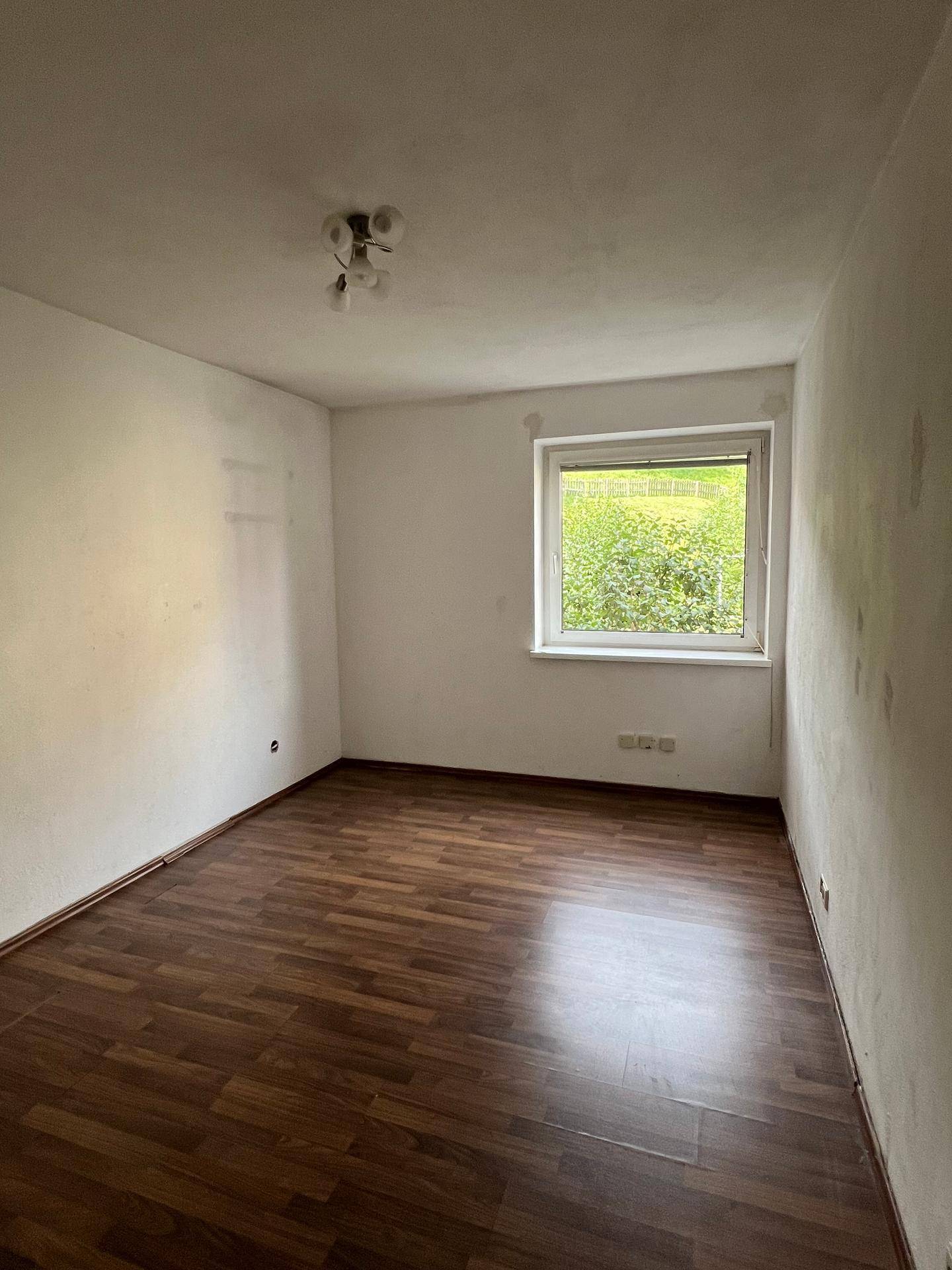 Schlafzimmer mit dunklem Laminatboden und Fenster mit Blick ins Grüne.