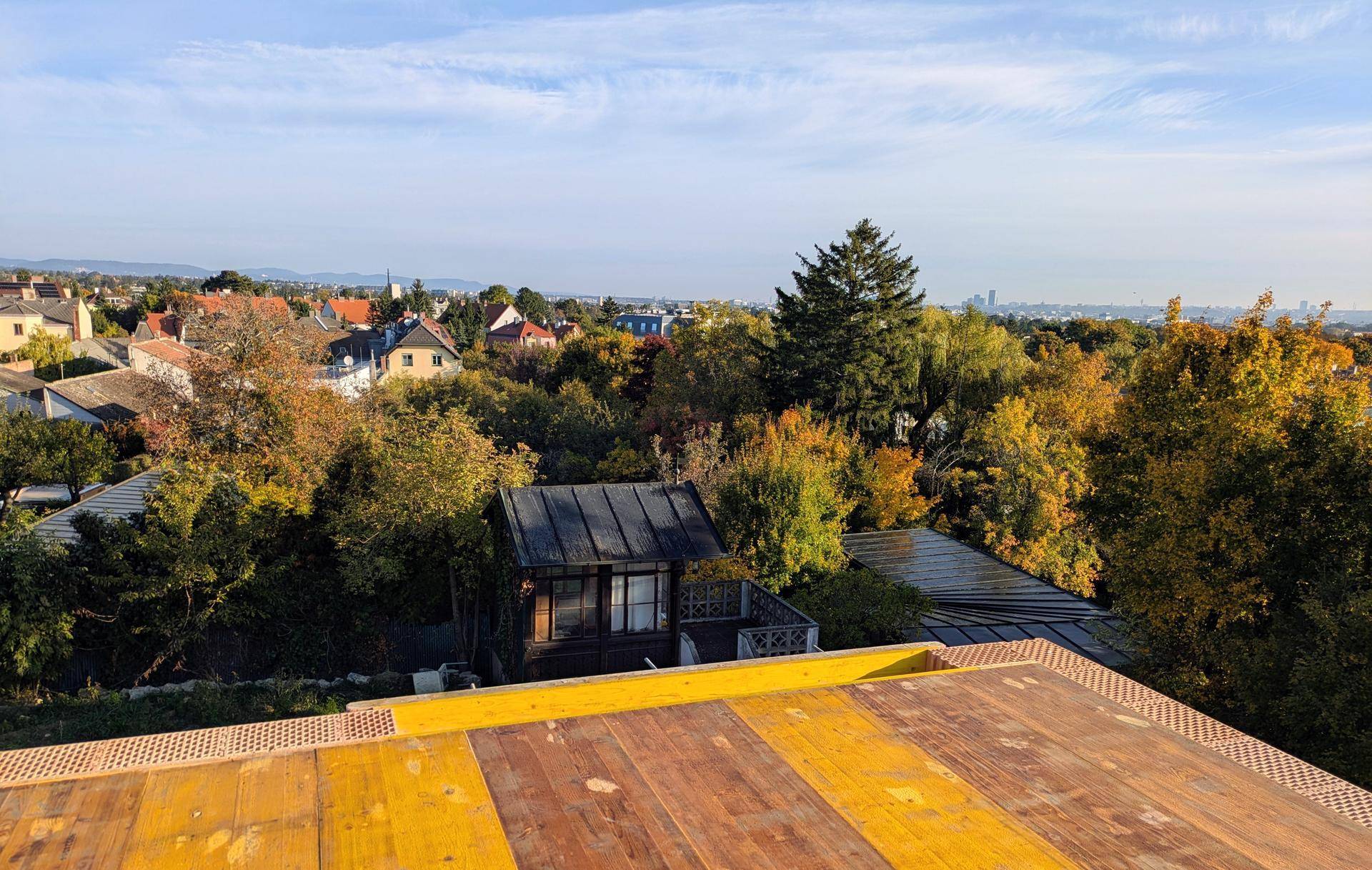 Weitläufiger Blick über eine grüne Wohngegend mit herbstlichen Bäumen und Stadtsilhouette am Horizont.