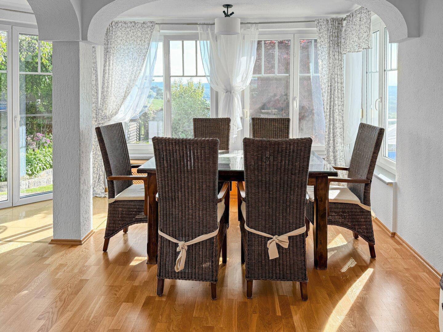 Heller Essbereich mit Rattan-Stühlen und Blick ins Grüne durch große Fenster.