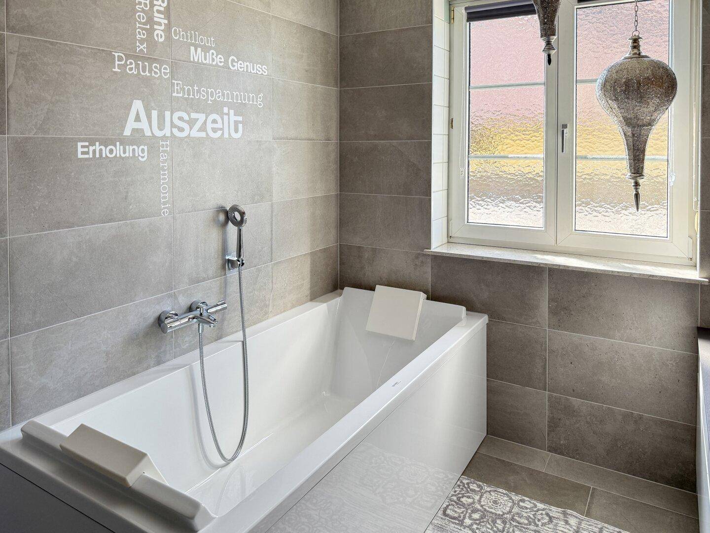Modernes Badezimmer mit Badewanne, grauen Fliesen und Fenster.