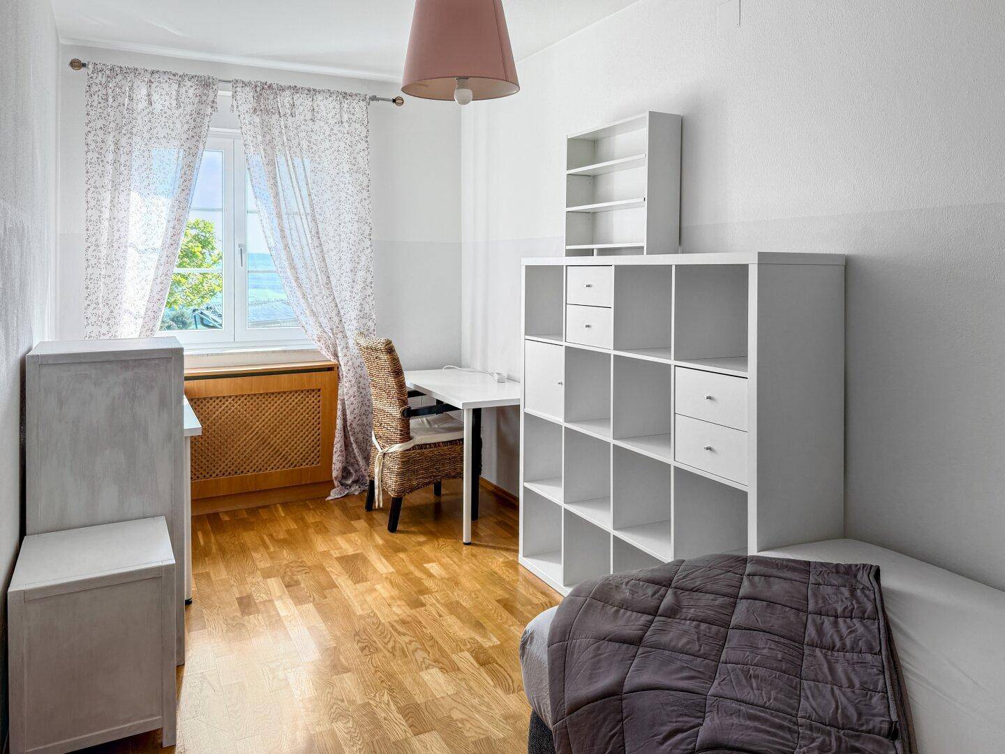 Helles Schlafzimmer mit Holzboden, Schreibtisch und Blick ins Grüne durch das Fenster.