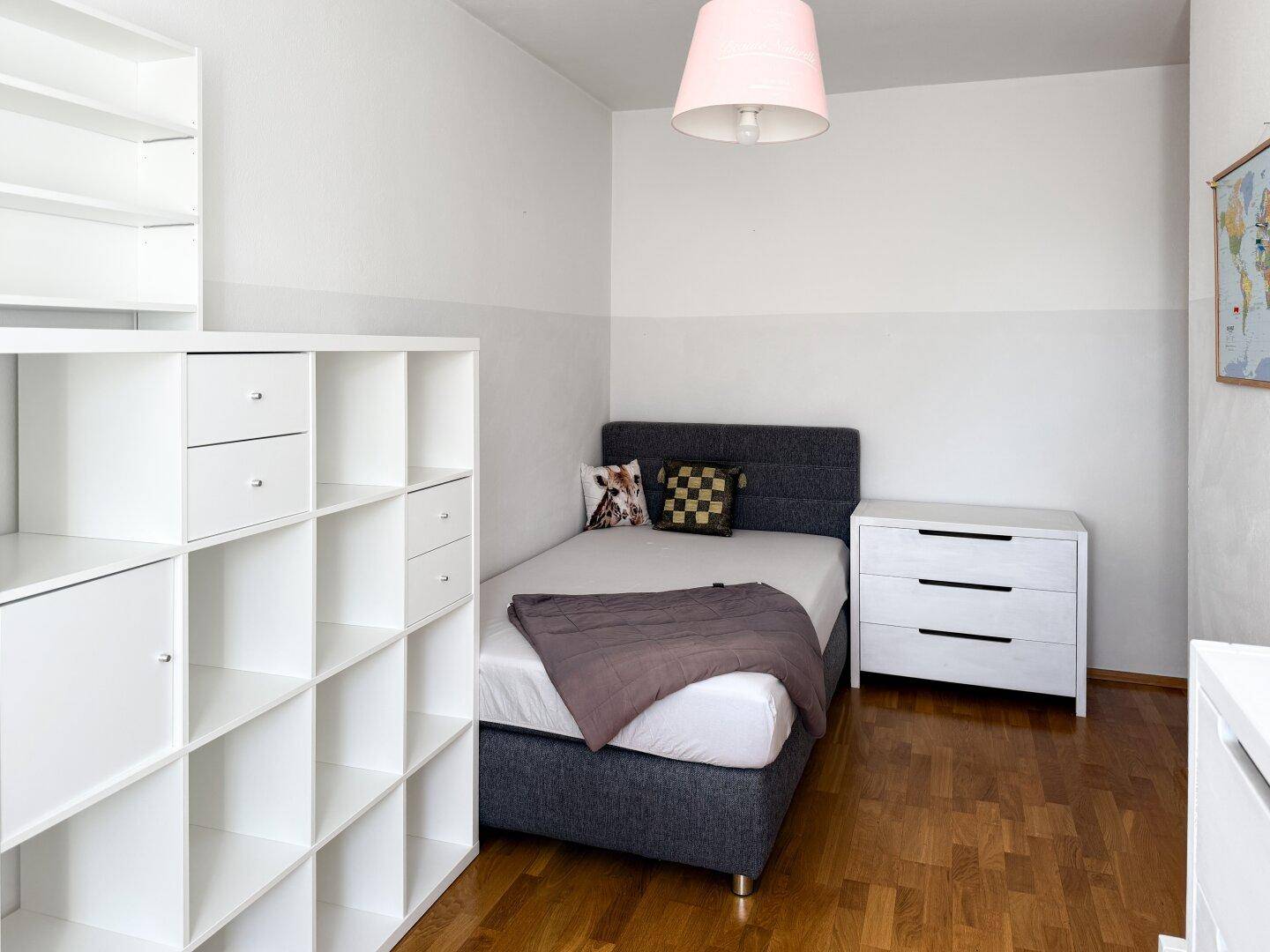 Modern eingerichtetes Schlafzimmer mit Einzelbett, Kommode und weißem Regalsystem.