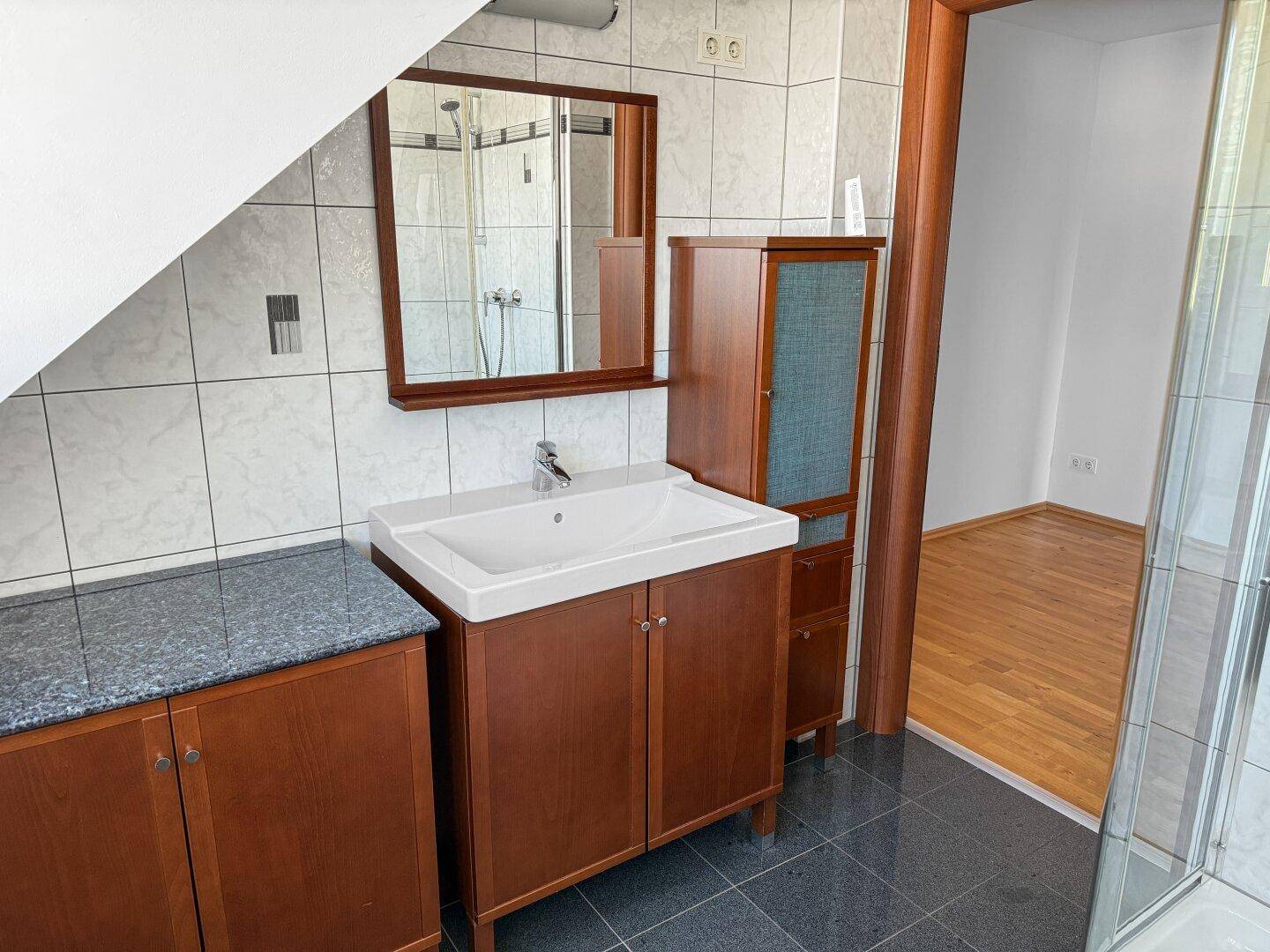 Badezimmer mit Waschtisch, Spiegel und dunklen Fliesen unter Dachschräge.