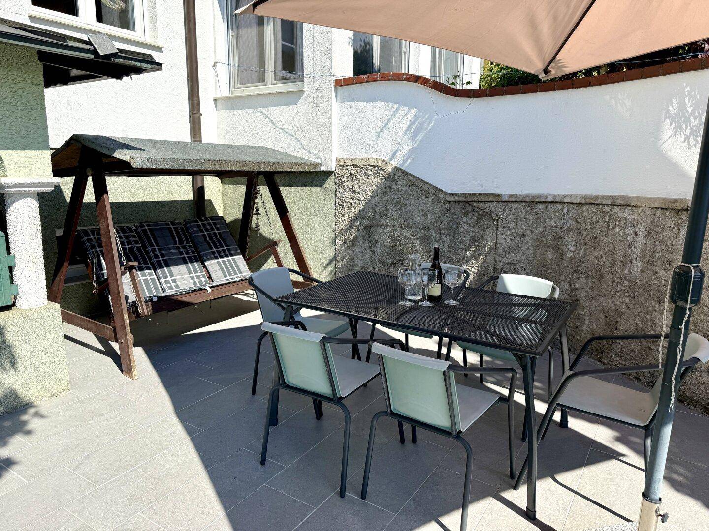 Gemütliche Terrasse mit Esstisch, Stühlen und einer Schaukelbank unter einem Sonnenschirm.