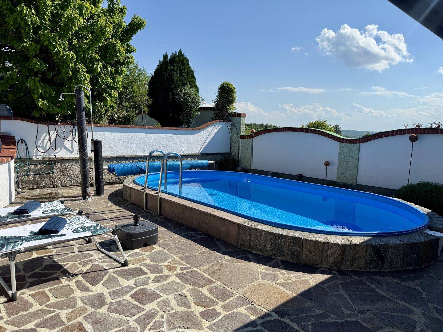 Ovaler Swimmingpool im Garten mit gepflasterter Liegefläche und Sonnenliegen.