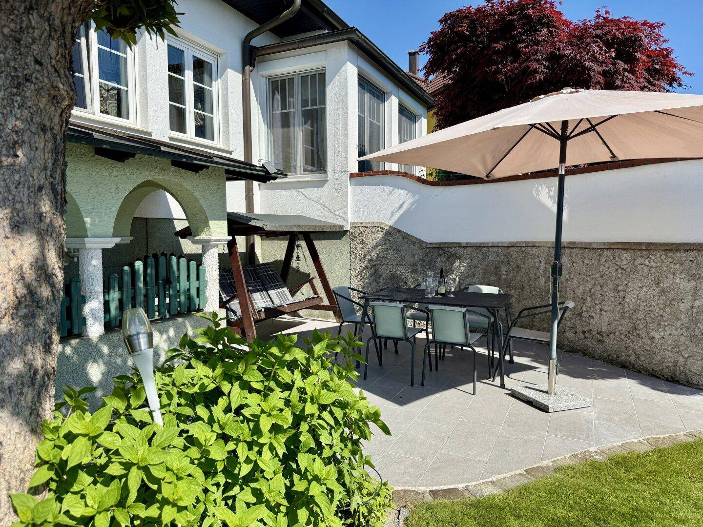 Helle Terrasse mit Esstisch, Stühlen und Schaukelbank, umgeben von üppigem Grün.