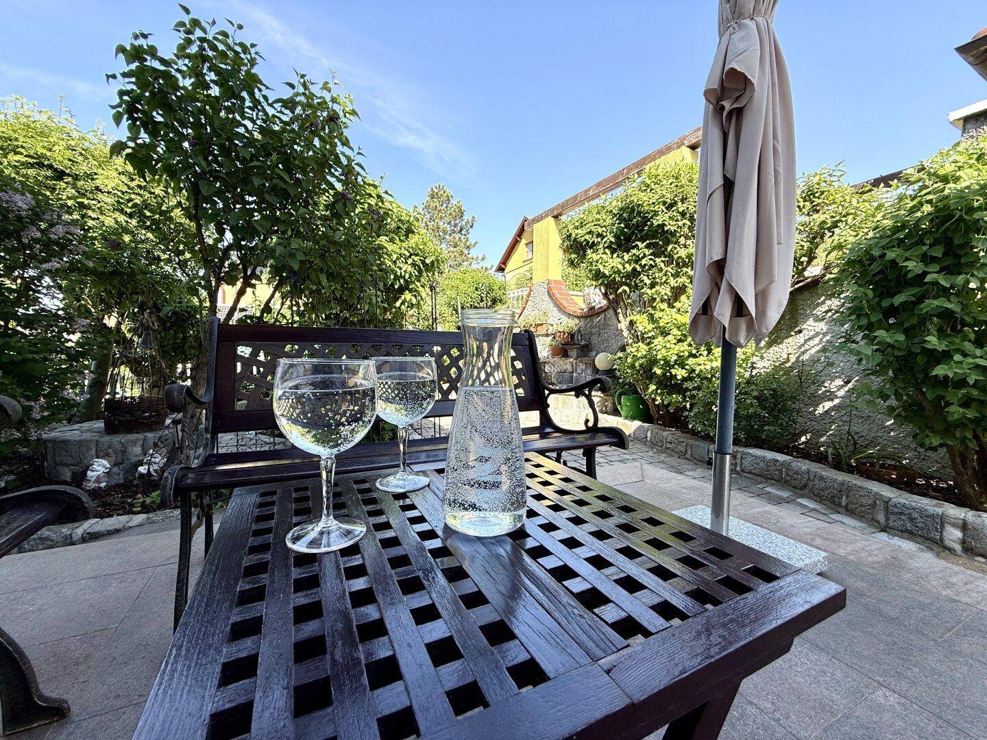 Gemütlicher Sitzbereich im Freien mit Holztisch und Bänken unter einem Sonnenschirm.