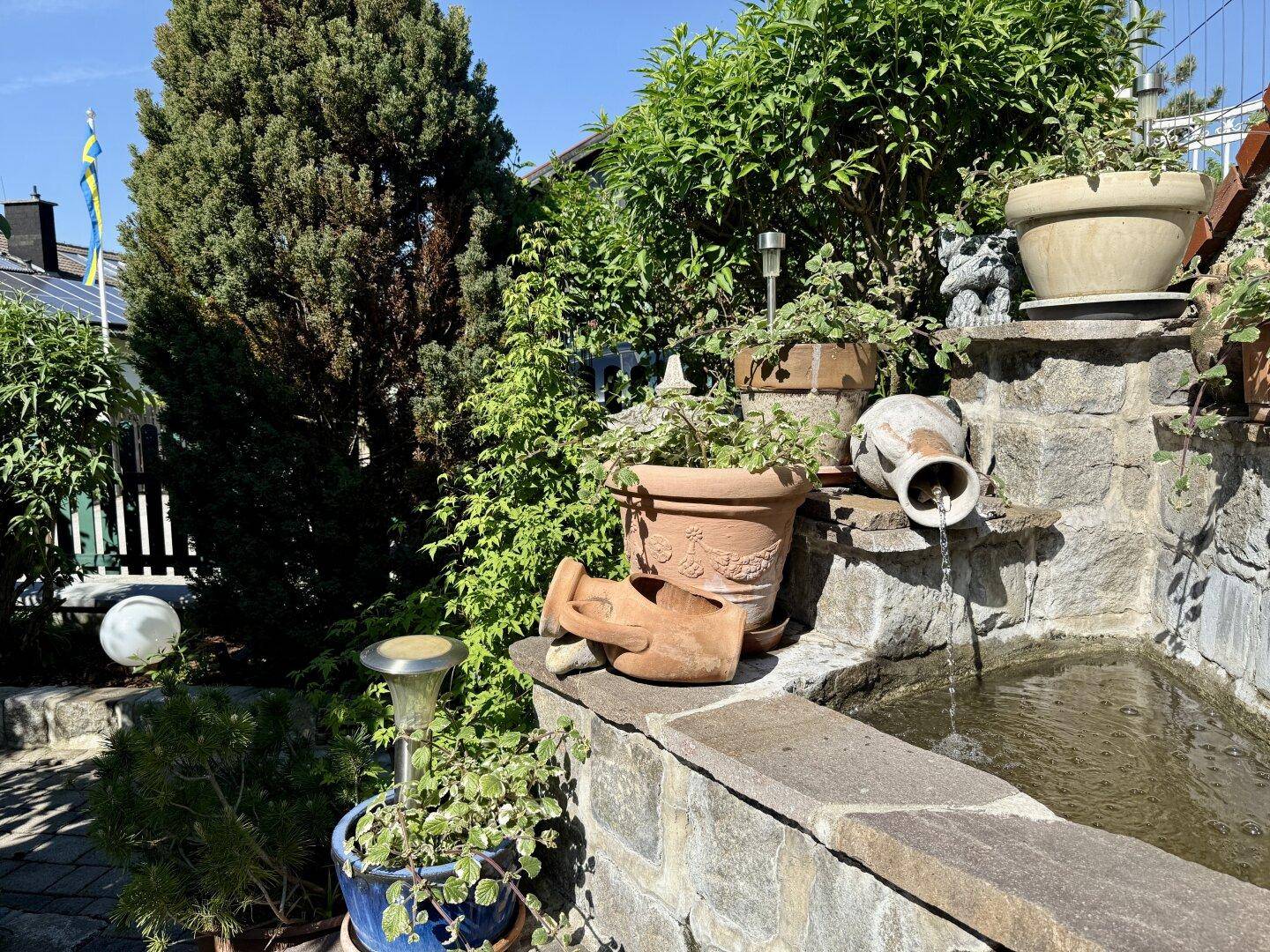 Dekorativer Gartenbrunnen aus Stein mit Topfpflanzen und üppigem Grün.