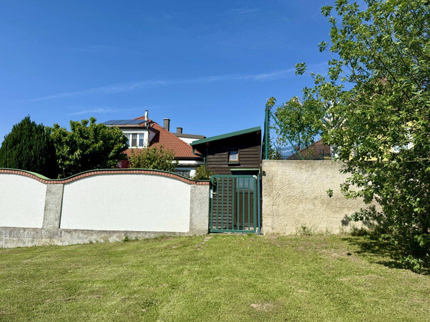 Grüner Zaun und Tor führen zu einem Teil des Hauses, umgeben von einer weißen Mauer und Bäumen.
