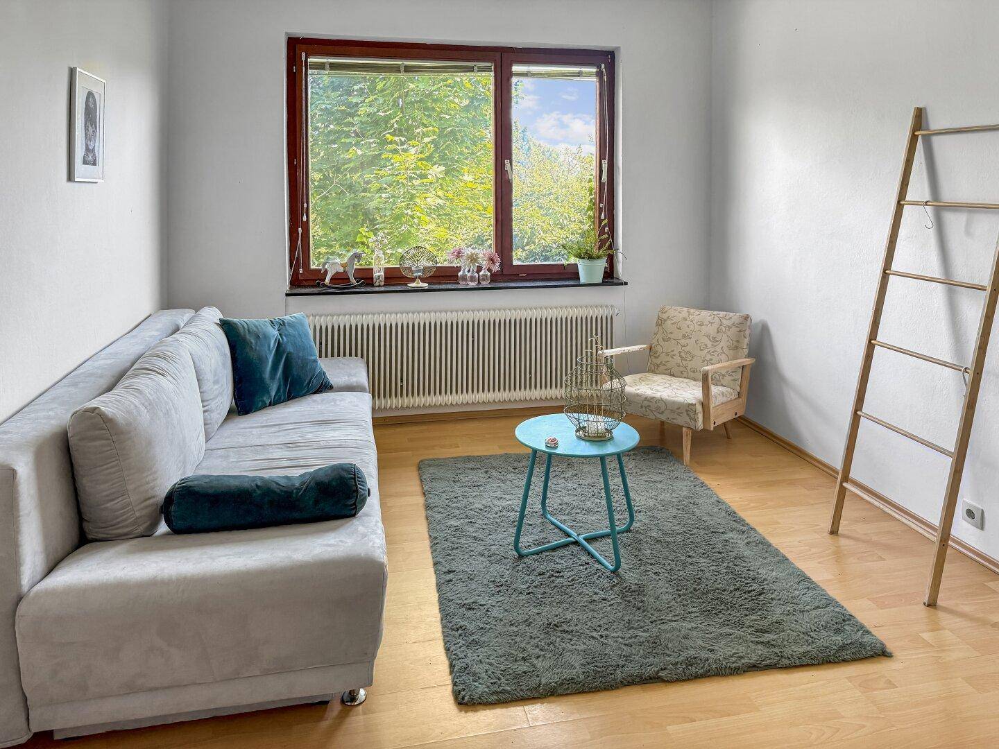 Gemütlicher Wohnbereich mit Sofa, Sessel und Blick ins Grüne durch das Fenster.