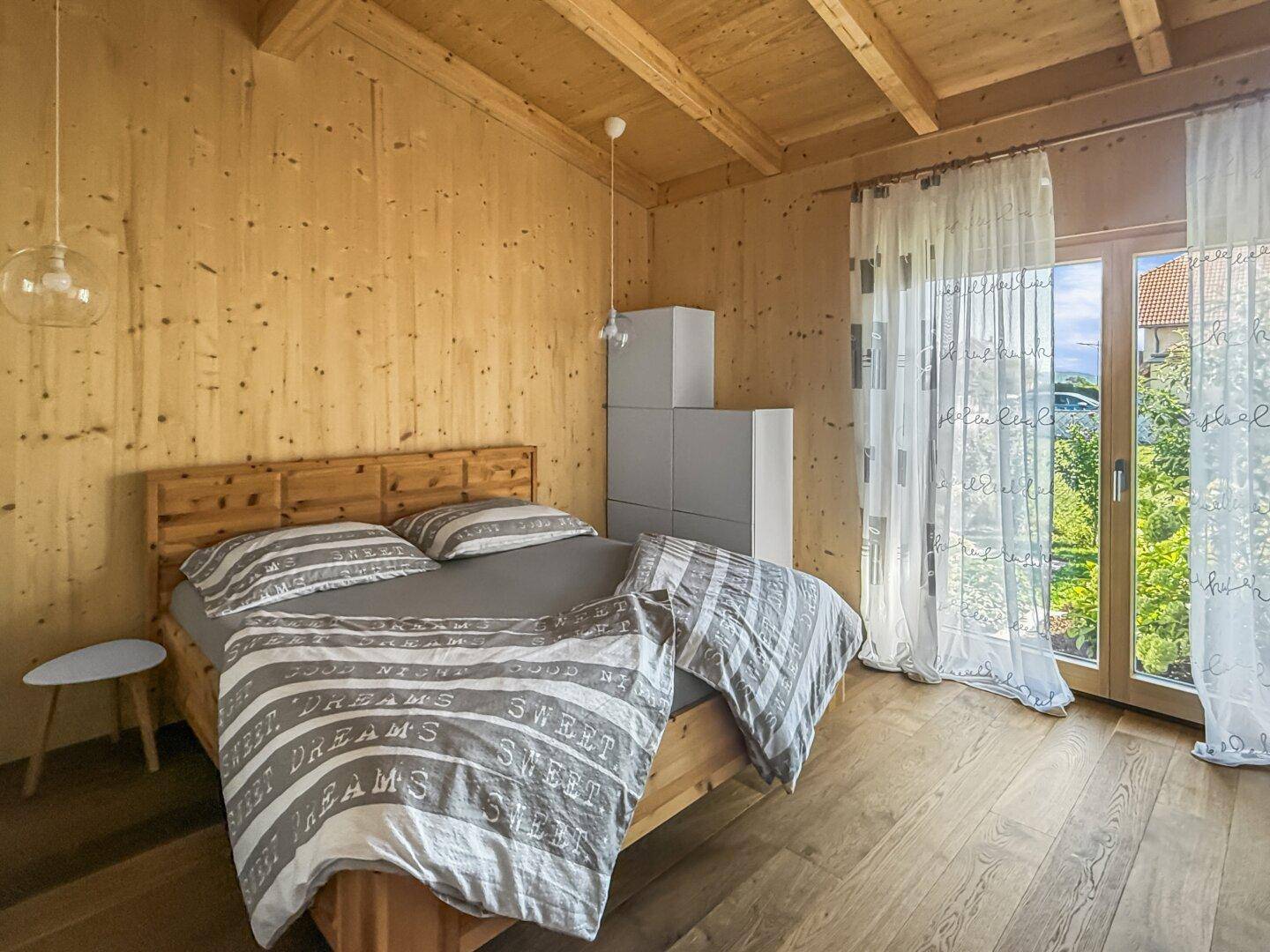 Helles Schlafzimmer mit Holzwänden, Holzbalkendecke und Zugang zum Außenbereich durch große Fenster.
