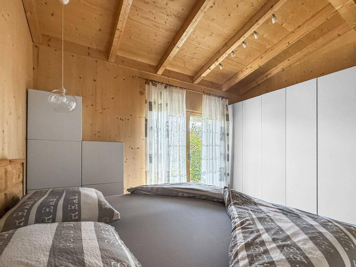 Schlafzimmer mit Holzbalkendecke, großen Fenstern und weißen Vorhängen, die viel Licht hereinlassen.