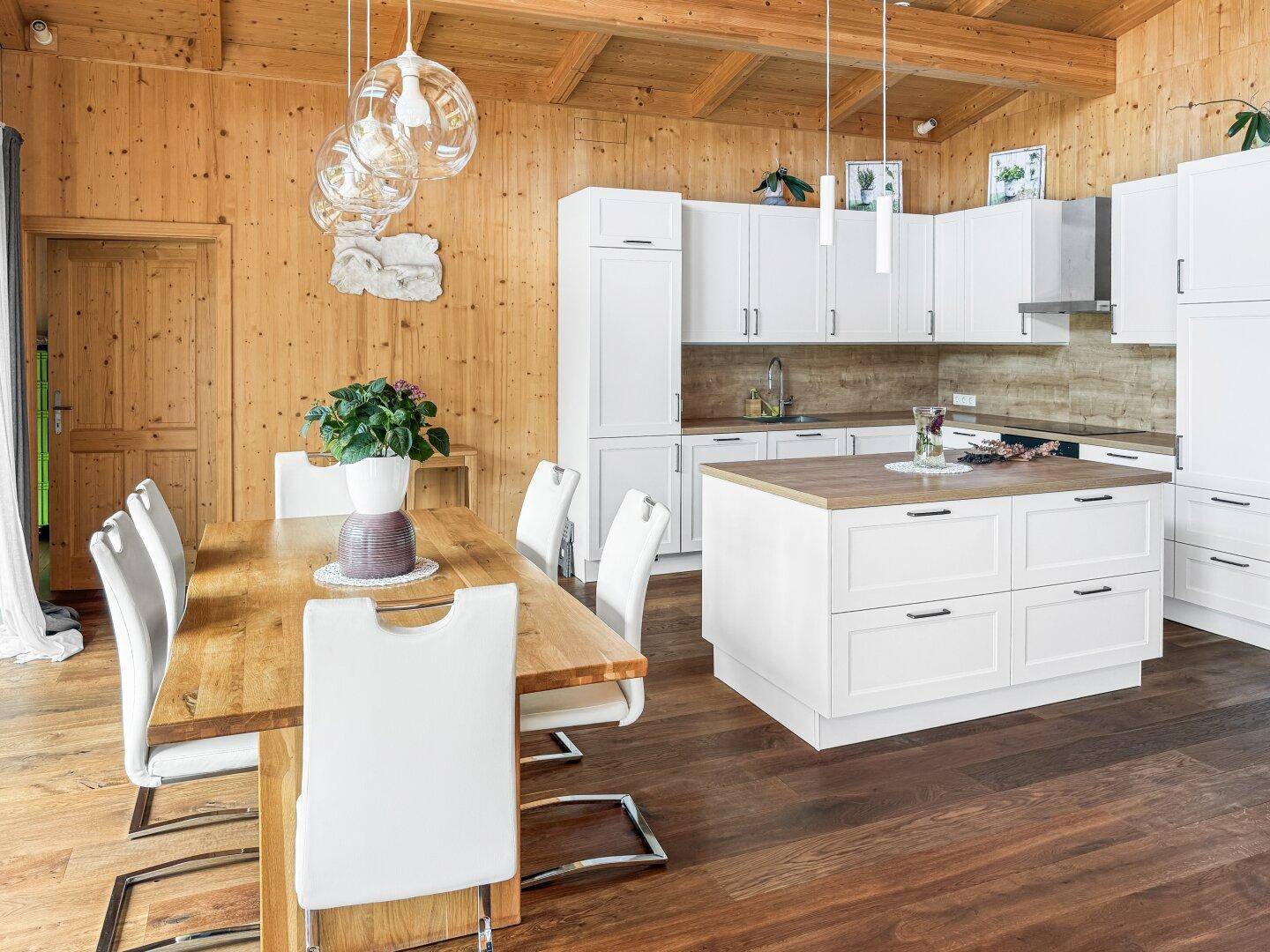 Offener Wohn-Essbereich mit moderner weißer Küche, Holztisch und Holzbalkendecke, die Gemütlichkeit ausstrahlt.