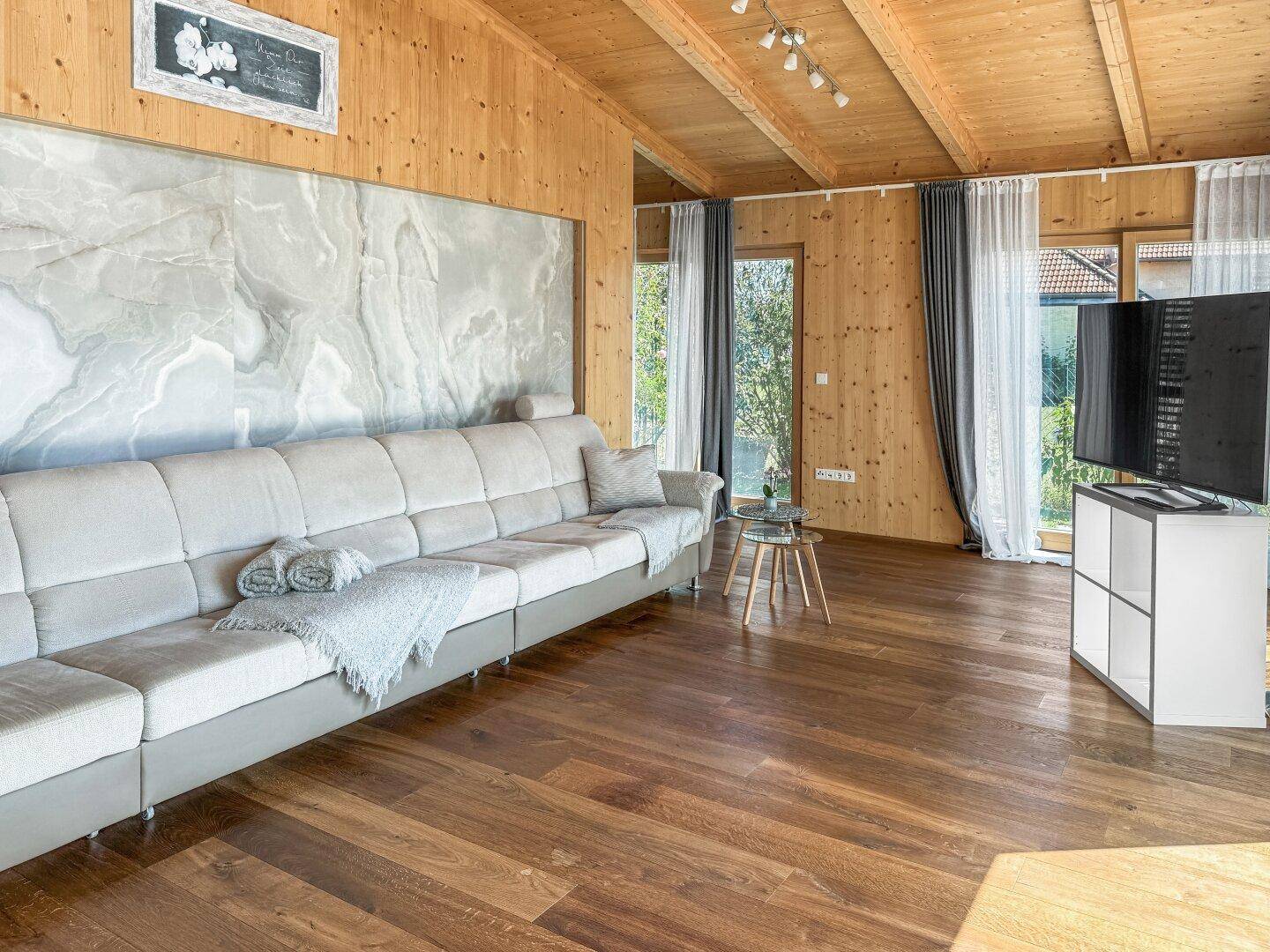 Gemütliches Wohnzimmer mit Sofa, Fernseher und viel Tageslicht durch die großen Fenster.
