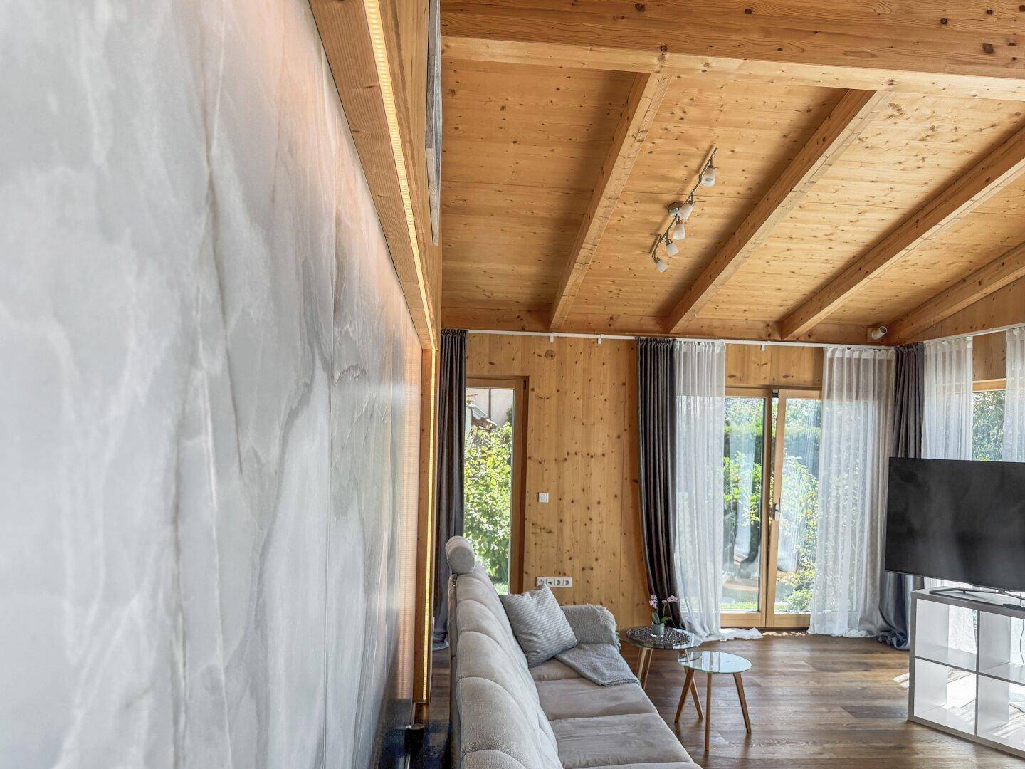 Moderner Wohnbereich mit beleuchteter Wandverkleidung, Holzbalkendecke und großen Fenstern.