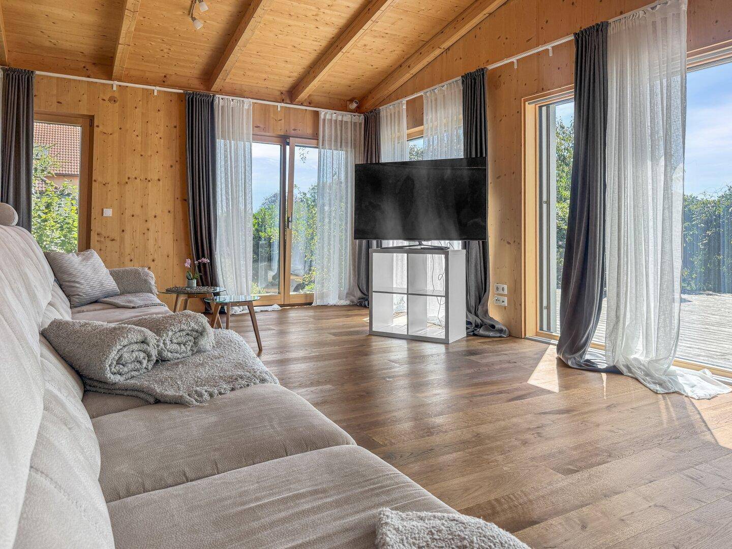 Heller Wohnbereich mit Fernseher, Holzboden und großen Fenstern, die den Raum erhellen.