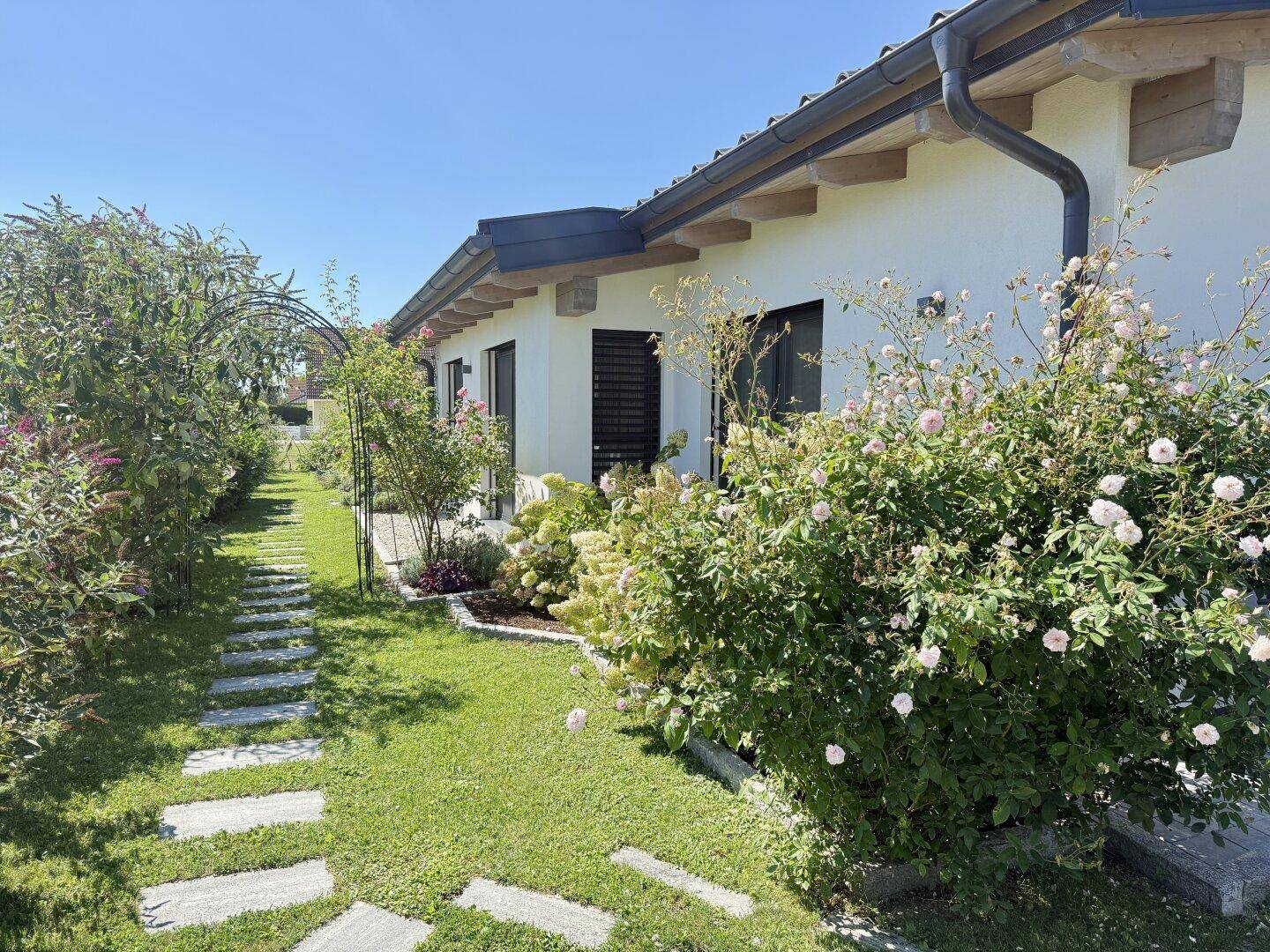 Seitlicher Gartenweg mit Rosenbüschen und einem Rankbogen, der entlang des Hauses verläuft.