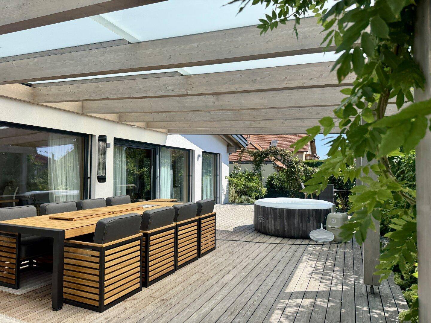Geräumige Holzterrasse mit überdachtem Essbereich und einem Whirlpool unter blauem Himmel.