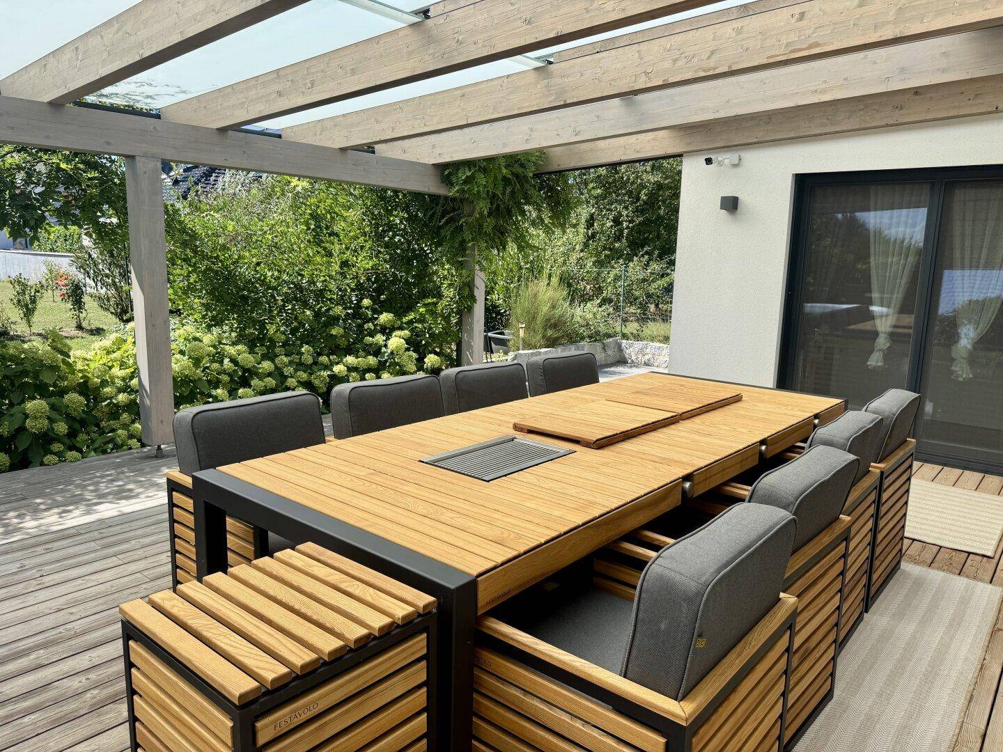 Überdachter Essbereich auf der Holzterrasse mit modernem Tisch und Stühlen, umgeben von Grün.