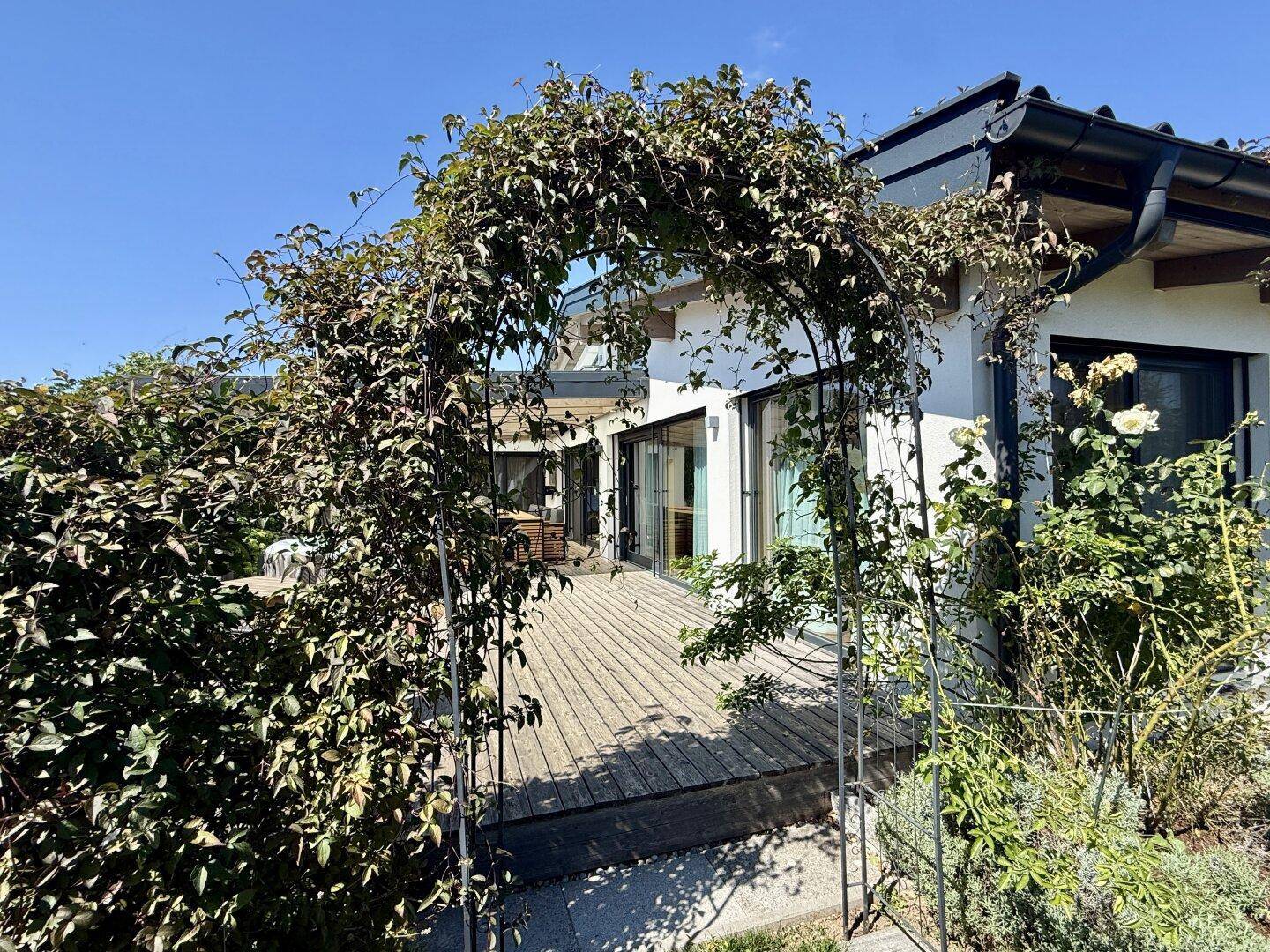 Gartenweg mit Rankbogen führt zu einer Holzterrasse am modernen Haus.