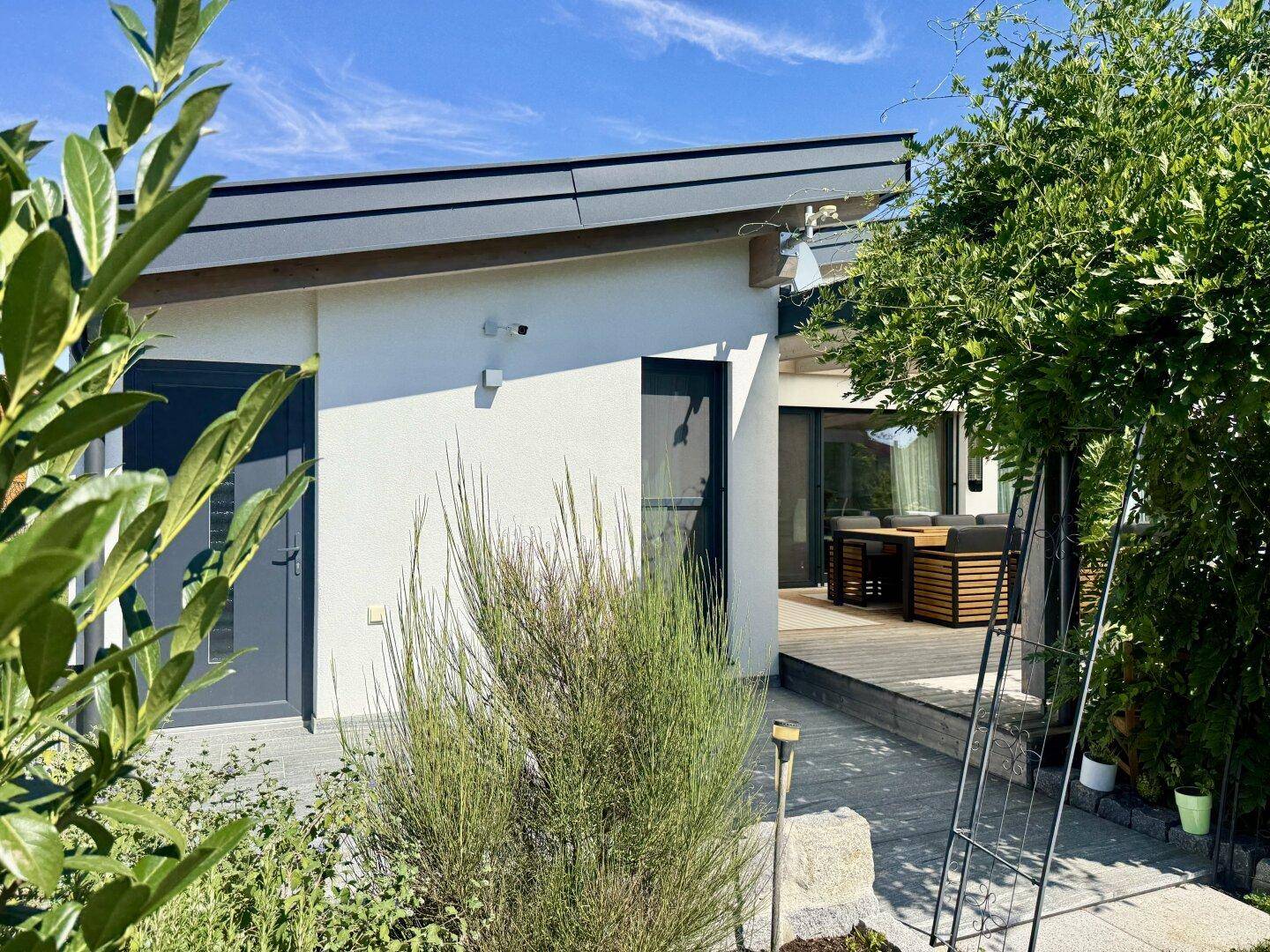 Seitlicher Gartenbereich mit Sträuchern und Blick auf die Holzterrasse und das moderne Haus.