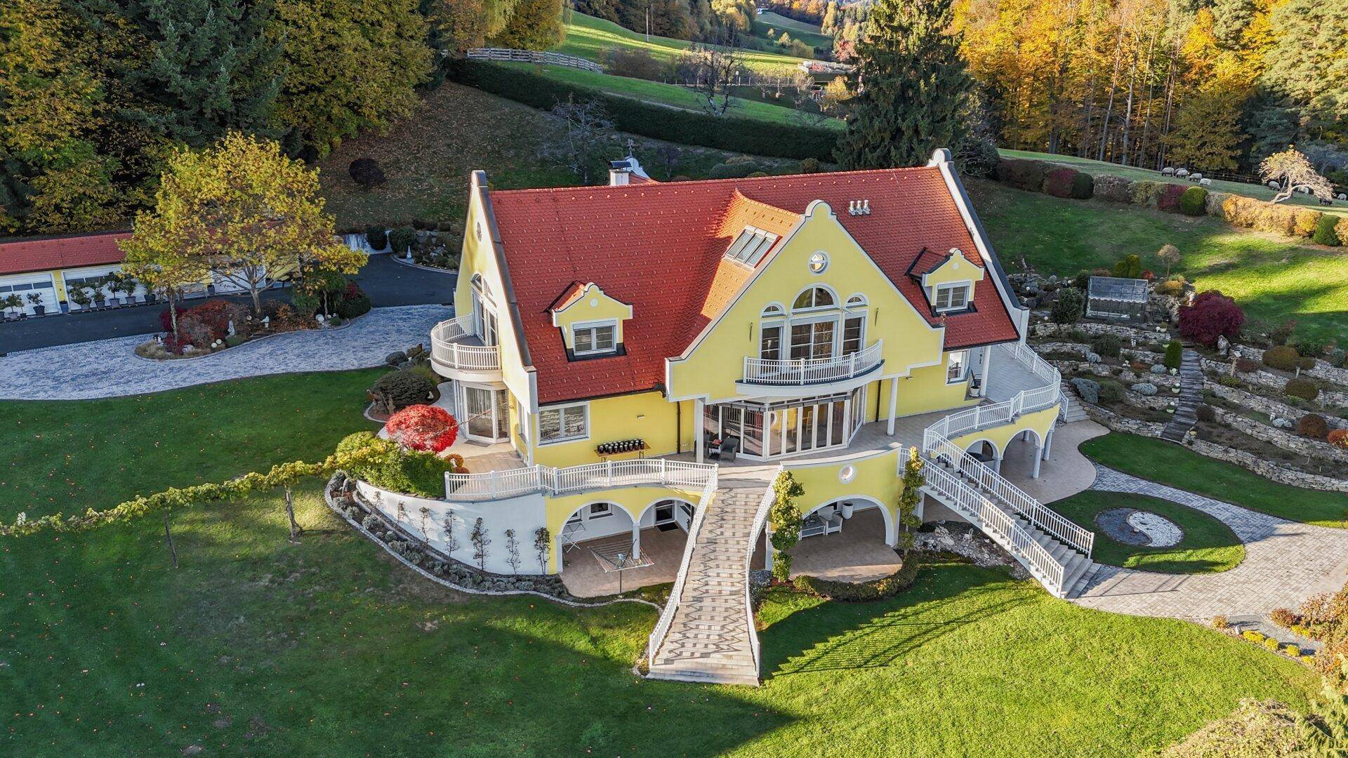 Luftaufnahme der gelben Villa mit rotem Dach, großem Garten und umliegender Hügellandschaft.