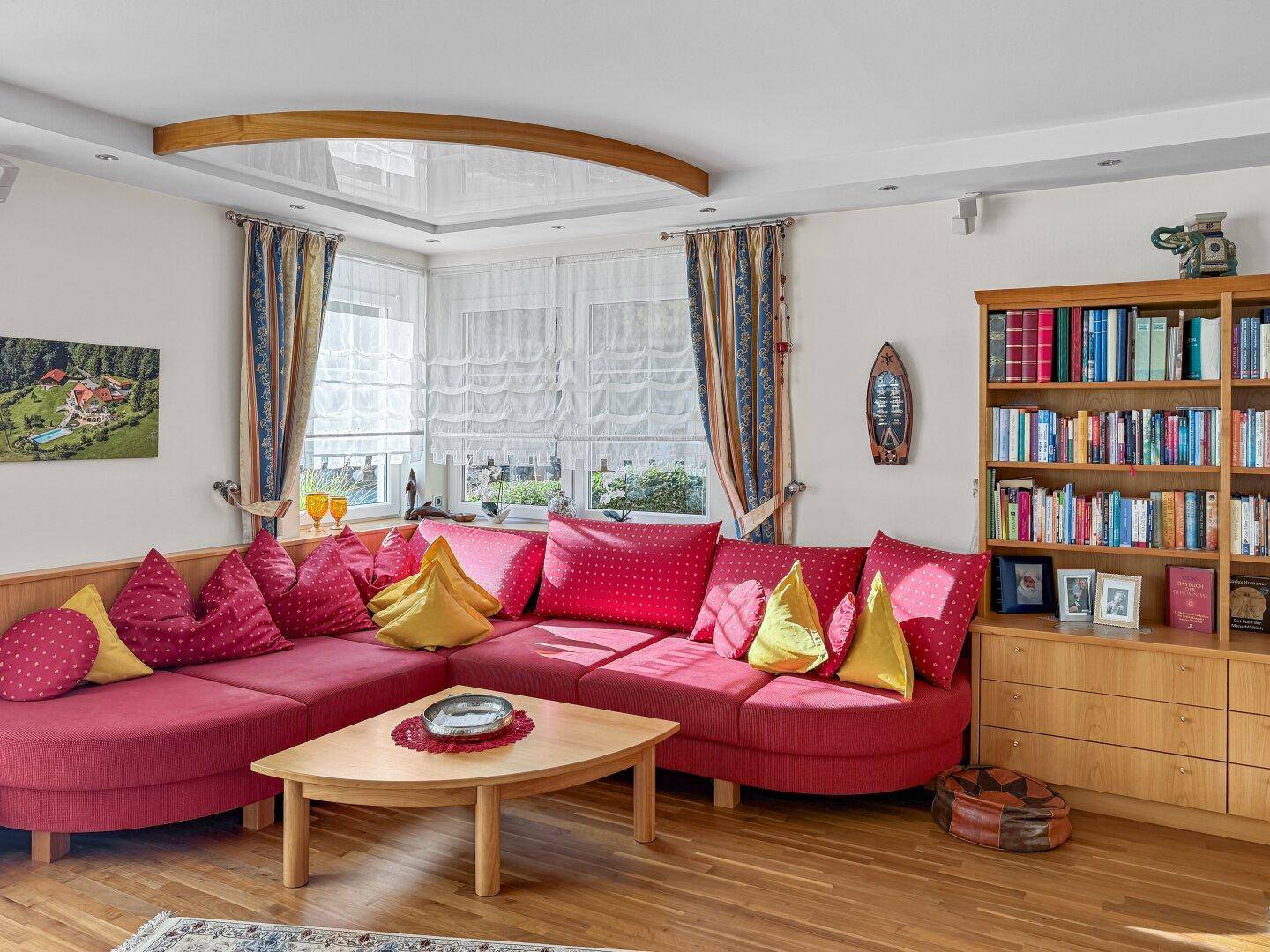 Farbenfrohes Wohnzimmer mit rotem Ecksofa, Bücherregal und Erkerfenster.