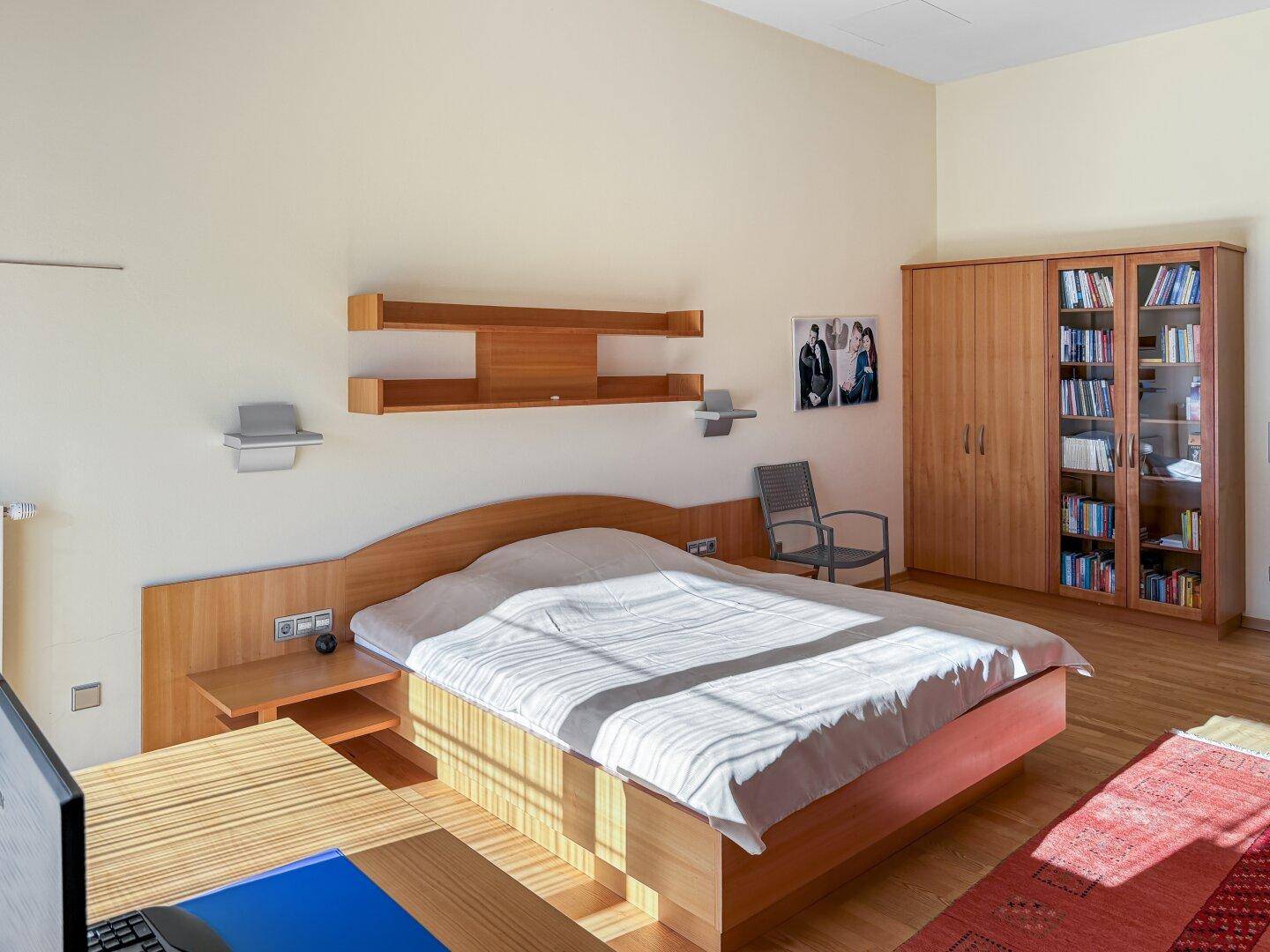 Schlafzimmer mit Holzbett, Schreibtisch und Bücherregal in hellem Ambiente.