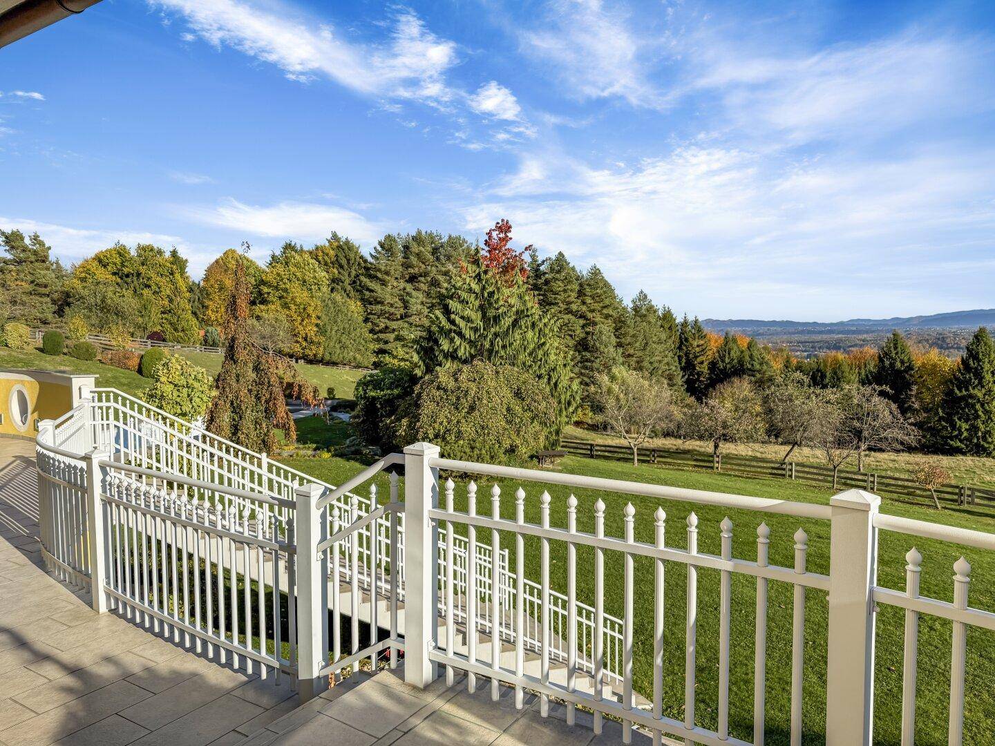 Terrasse mit weißem Geländer und Treppe, die zu einem weitläufigen Garten mit Fernblick führt.