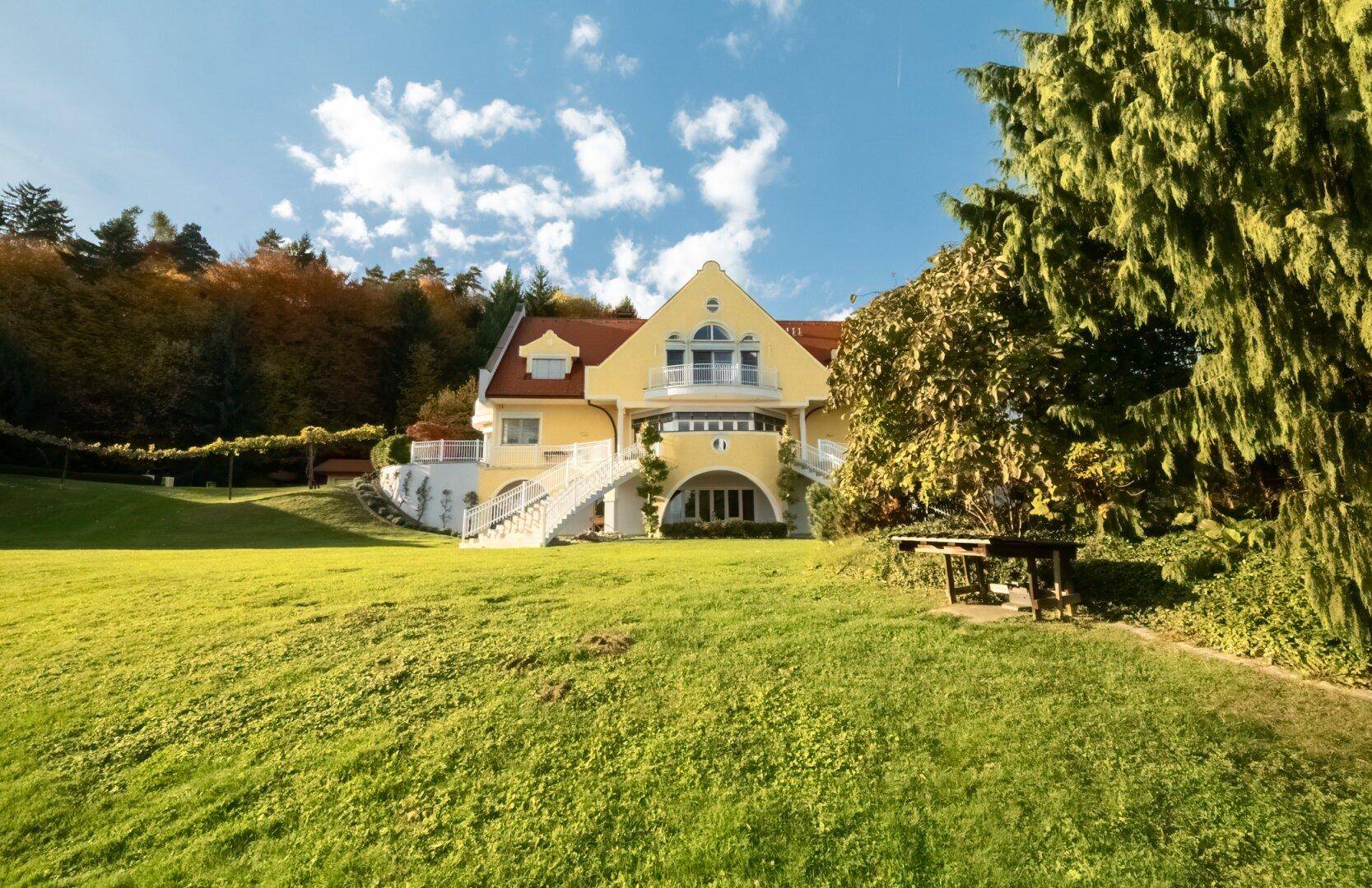 Große gelbe Villa mit rotem Dach auf einem grünen Hügel, umgeben von Bäumen.