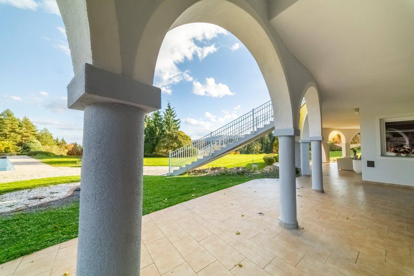 Blick durch einen Bogen auf eine geflieste Terrasse mit Säulen und einer Treppe zum Garten.