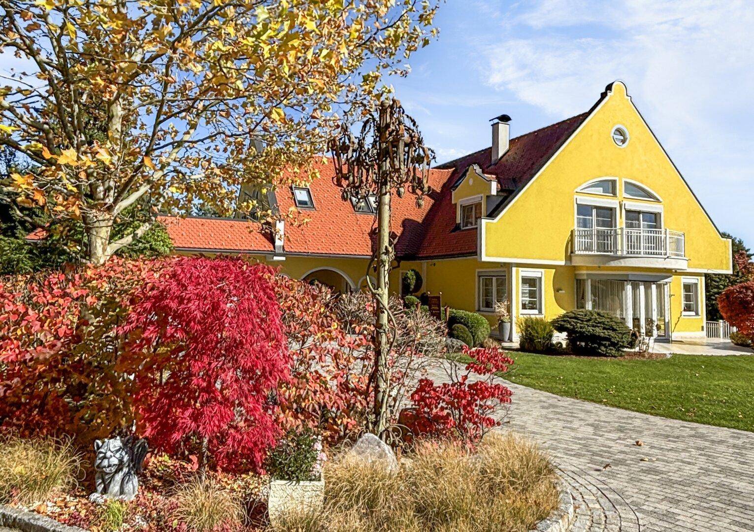 Gelbe Villa mit rotem Dach, umgeben von farbenfrohem Herbstlaub und gepflasterter Auffahrt.