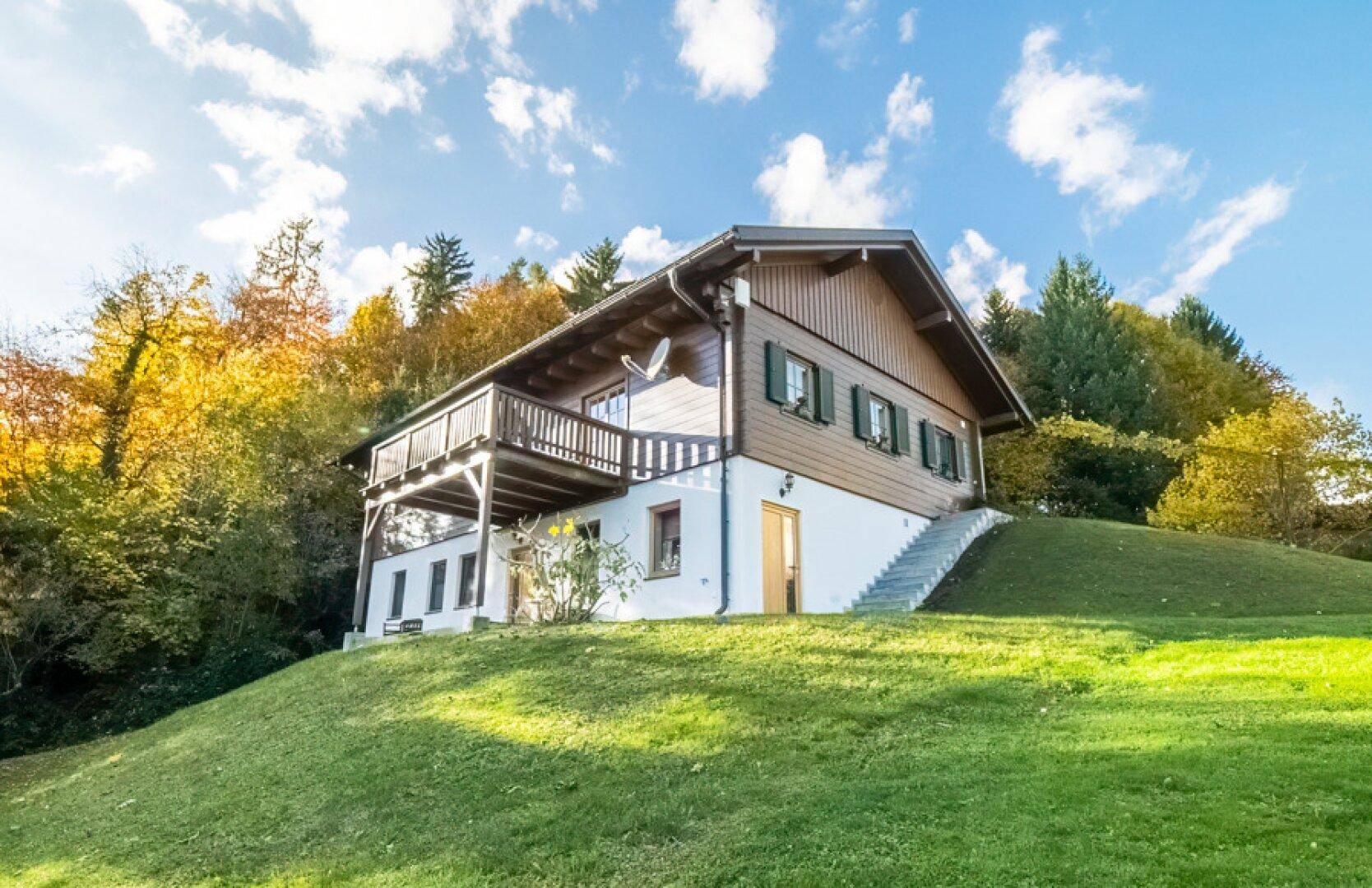 Traditionelles Holzhaus auf einem grünen Hügel, umgeben von herbstlichen Bäumen und Natur.