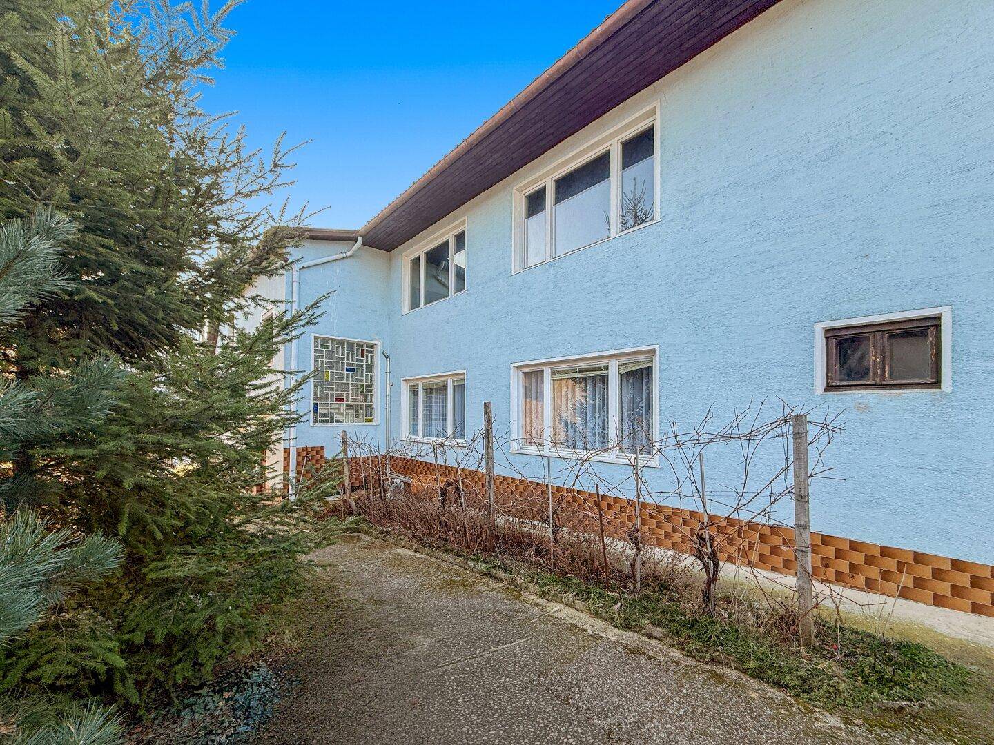 Seitenansicht des Hauses mit blauer Fassade, mehreren Fenstern und einem schmalen Weg.
