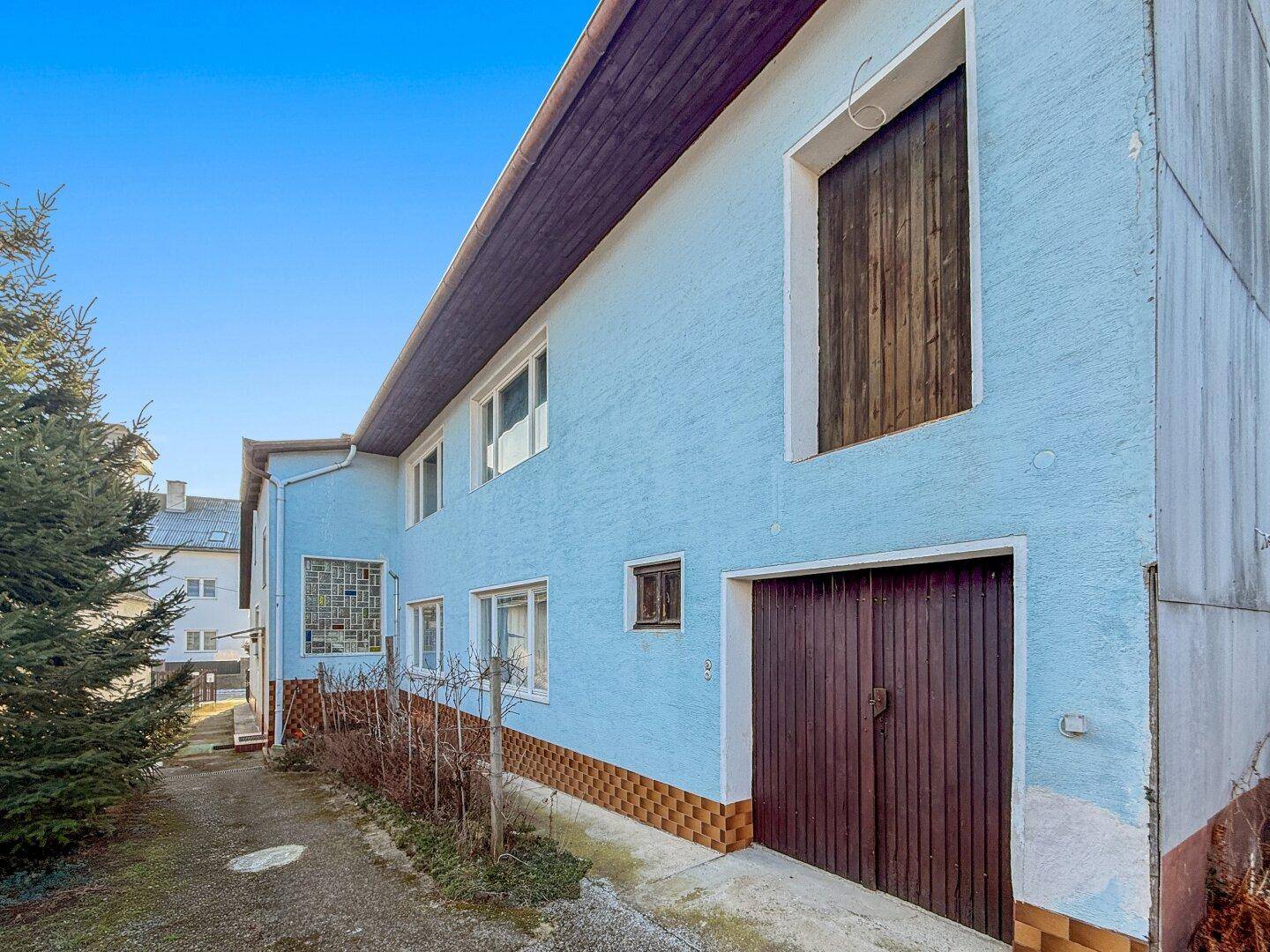 Seitenansicht des Hauses mit blauer Fassade und einer großen Holztür, möglicherweise ein Garagenzugang.