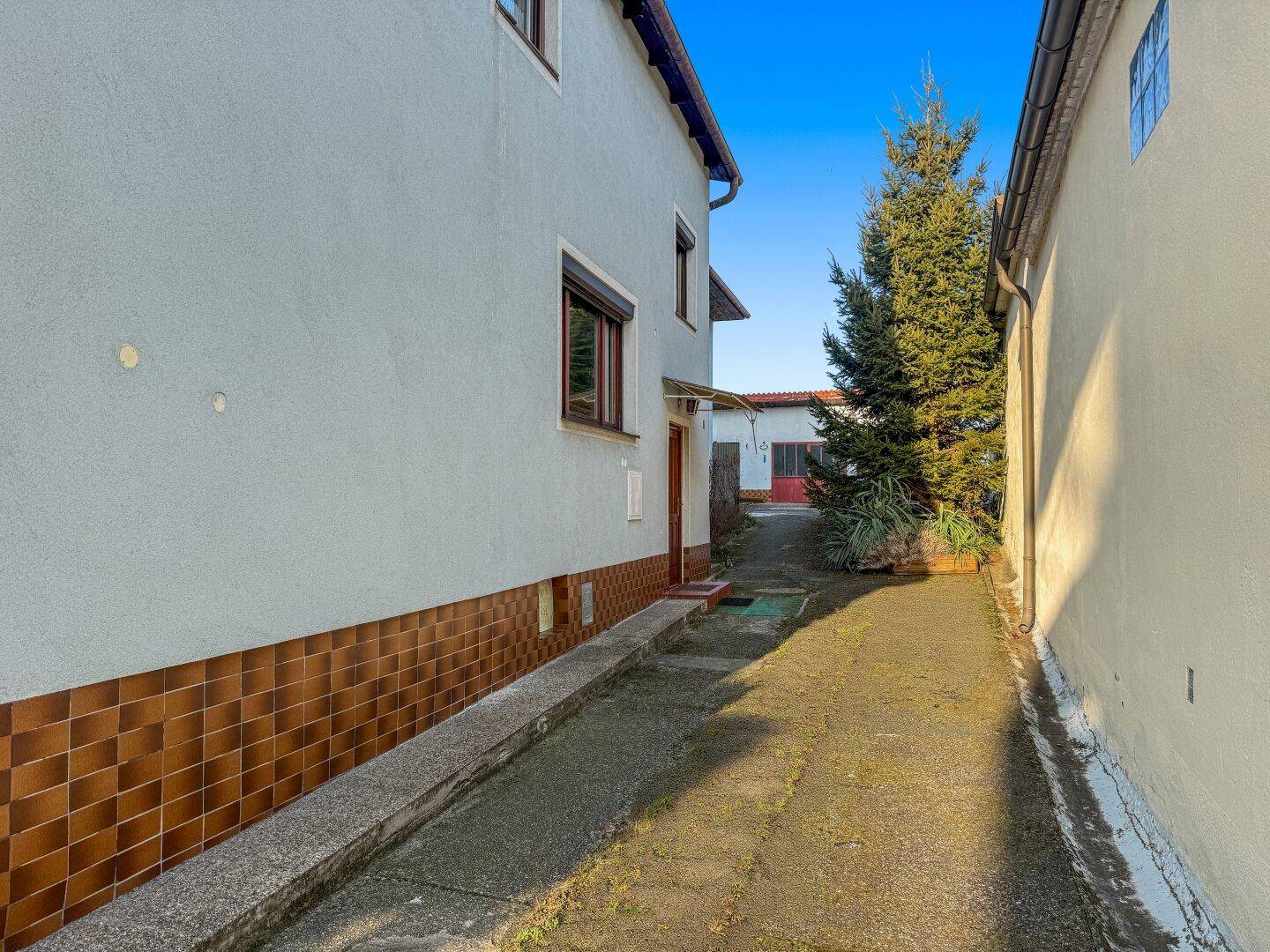 Schmaler Weg zwischen zwei Gebäuden mit gefliester Sockelzone und einem Nadelbaum.