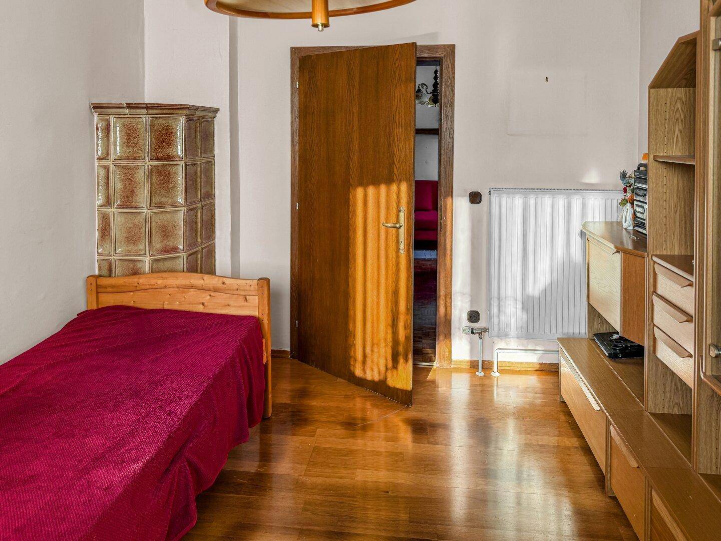 Helles Schlafzimmer mit Einzelbett, Holzschrank und einem Kachelofen im Hintergrund.