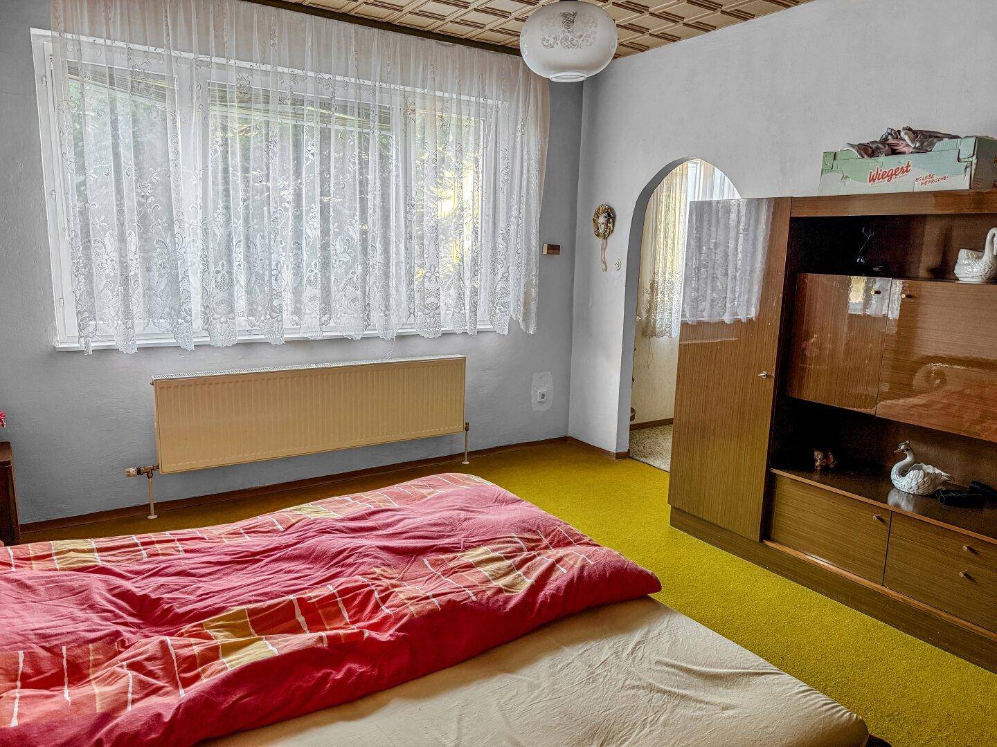 Schlafzimmer mit großem Fenster, Heizkörper und Blick auf den Garten.