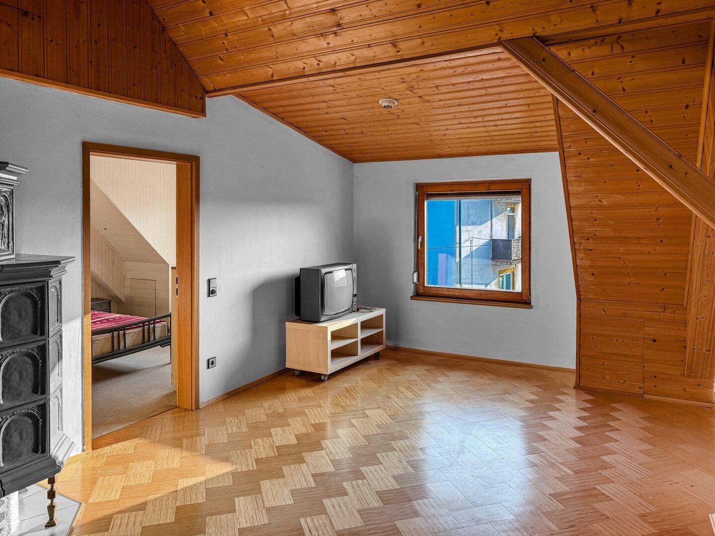 Dachgeschoss-Wohnzimmer mit Parkettboden, Holzbalkendecke und Fenster mit Blick ins Grüne.