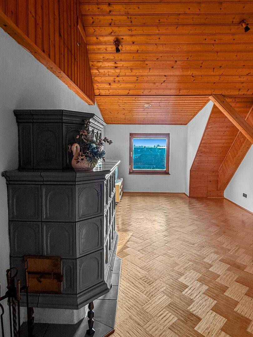Dachgeschoss-Wohnzimmer mit Parkettboden, Holzbalkendecke und einem dunklen Kachelofen.