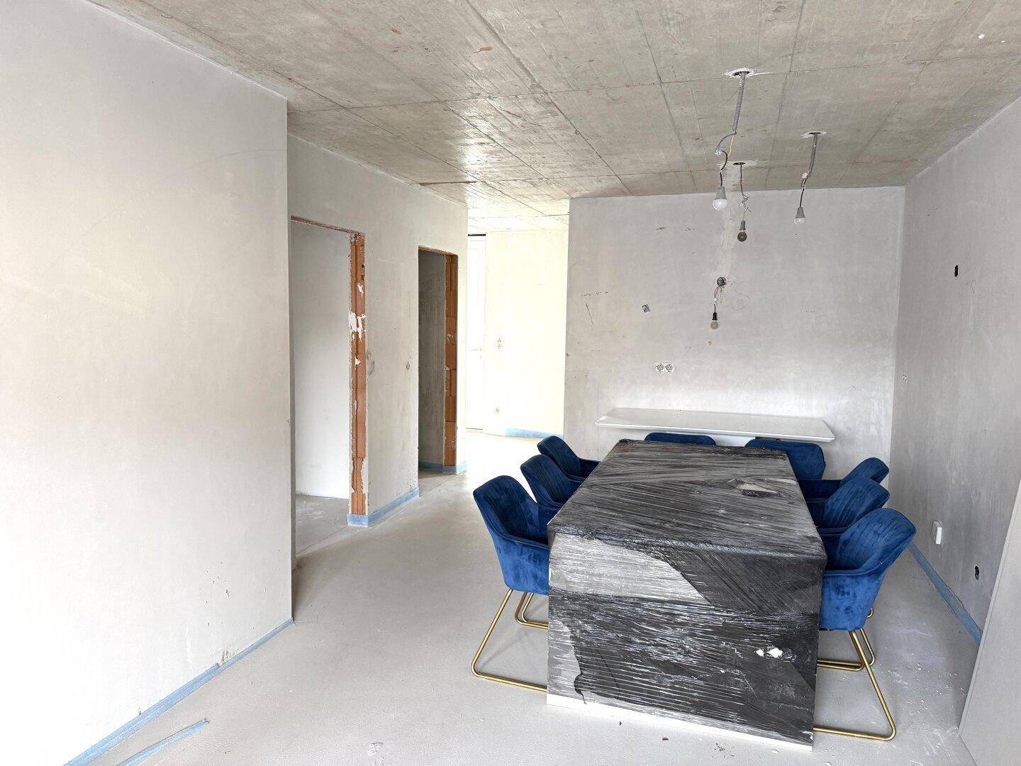 Essbereich im Rohbauzustand mit unfertigen Wänden und Decke, blauen Stühlen und verpacktem Tisch.